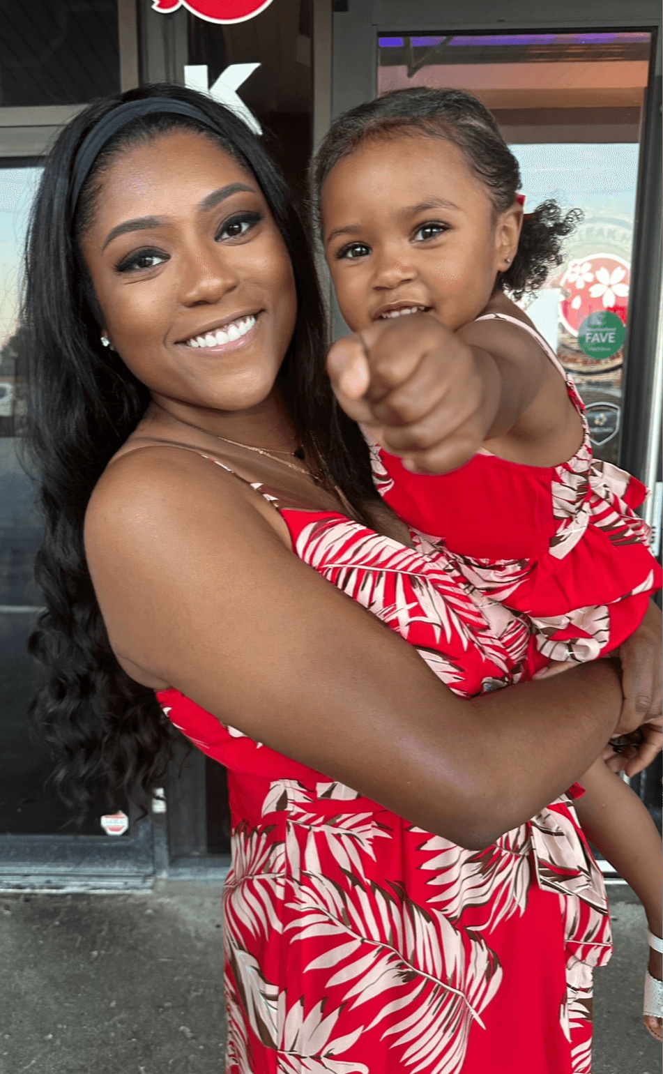 Smiling woman holding a young girl pointing at the camera indoors with a window behind them.