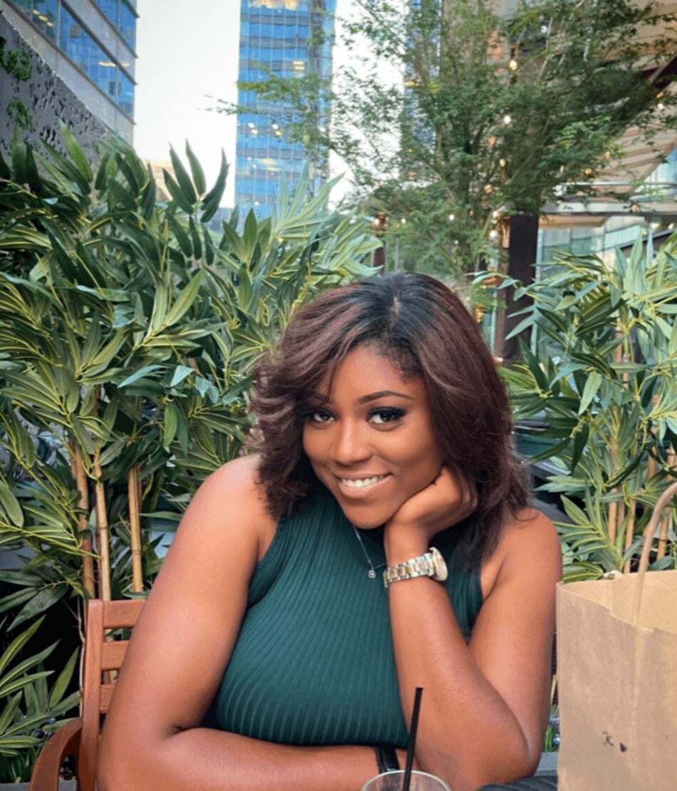 Smiling woman with shoulder-length wavy hair, wearing a sleeveless green top, sitting outdoors with greenery and buildings behind her.