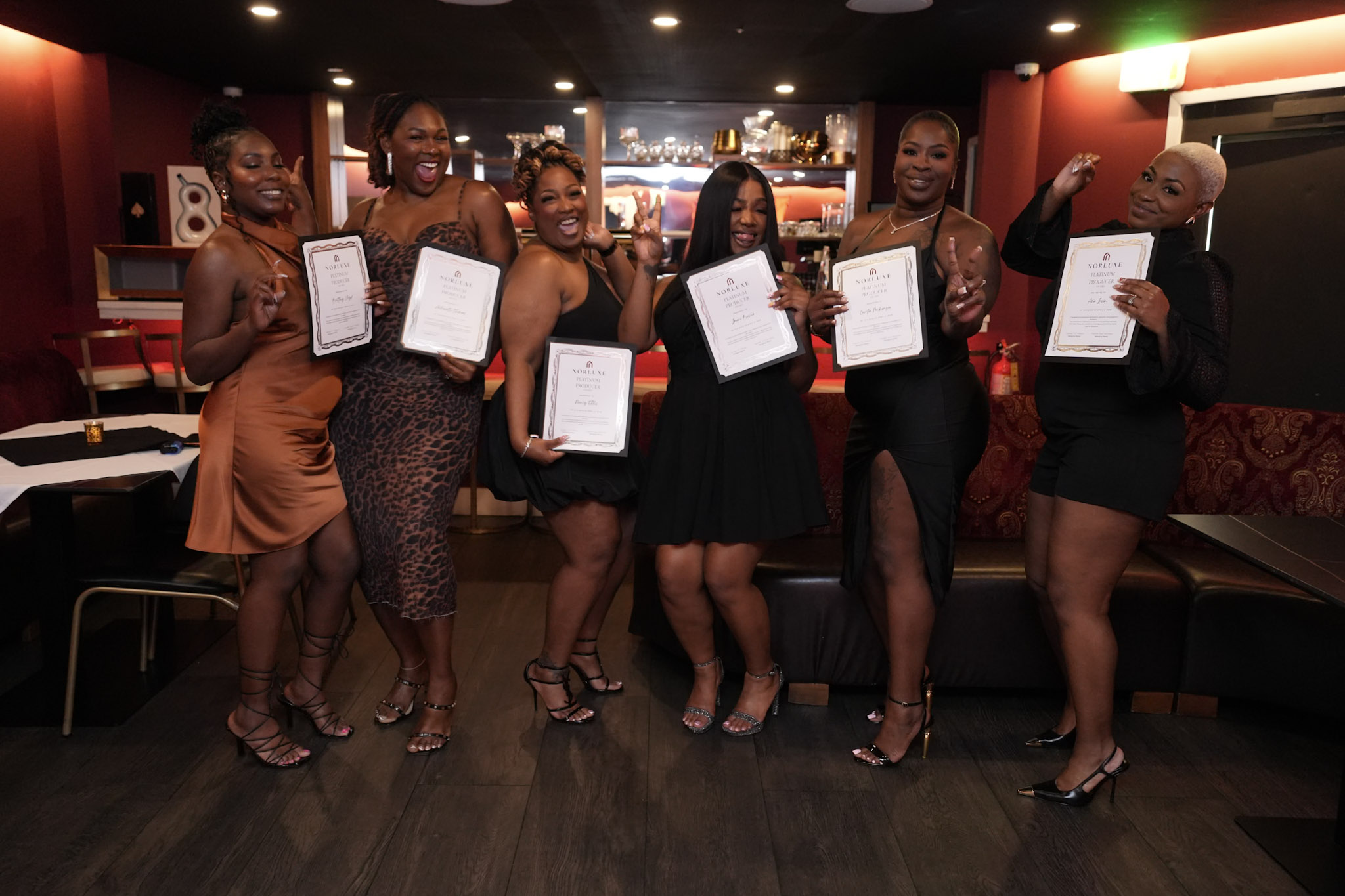 Seven women in dresses holding certificates, smiling in a dimly lit room with red walls and a bar in the background.