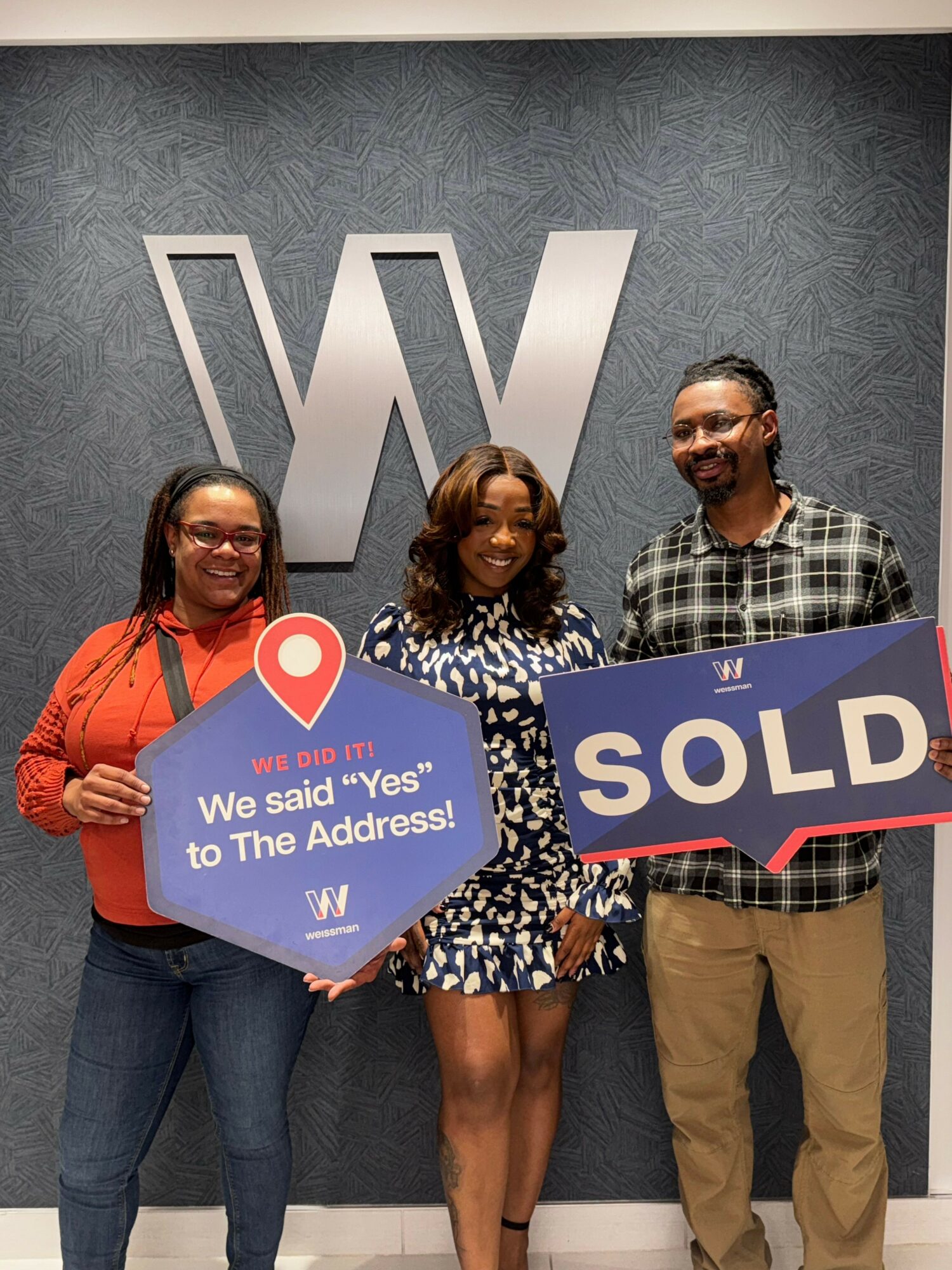 Three people holding signs in front of a wall with a large logo, celebrating a real estate sale.