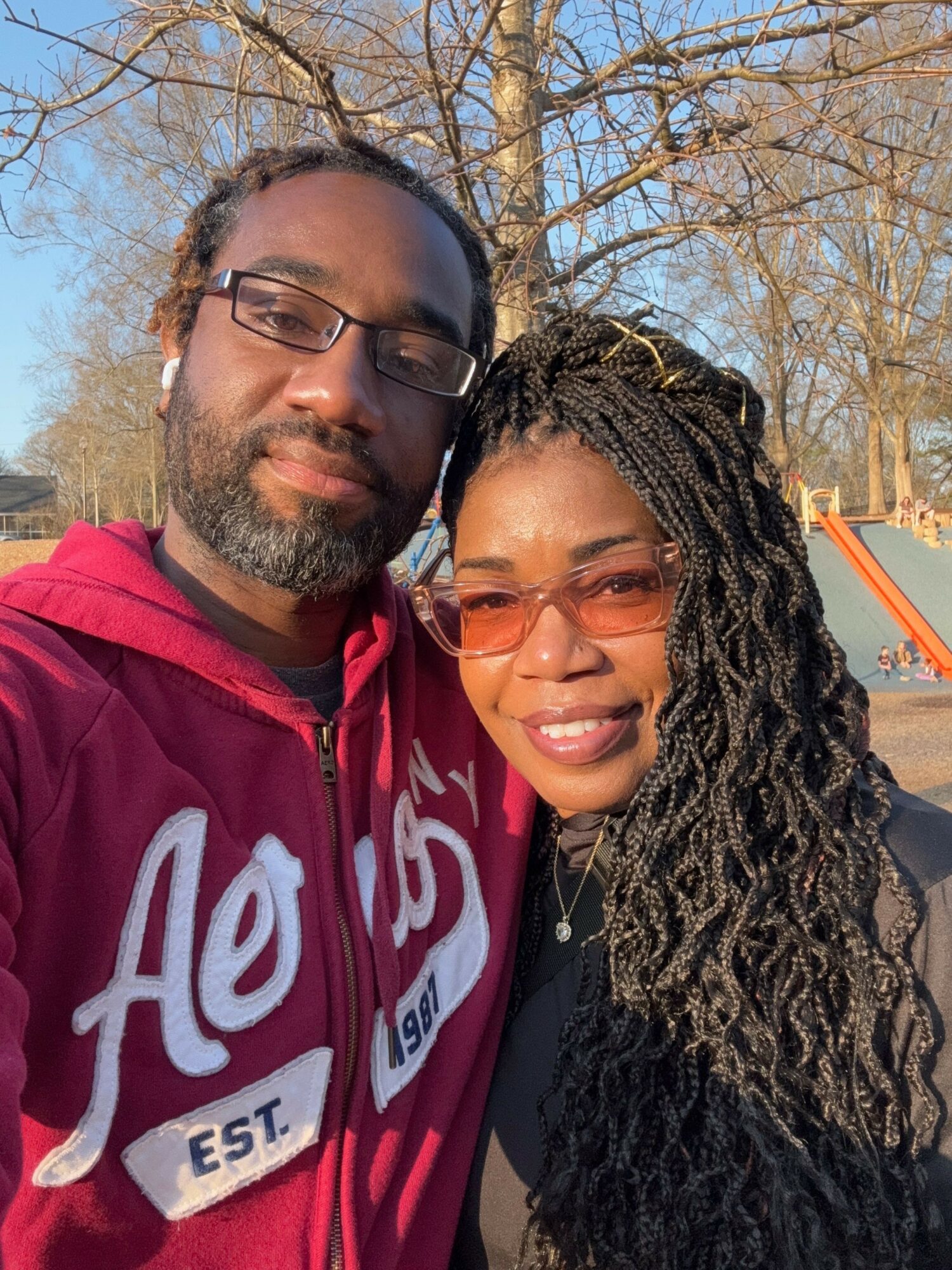 A man and woman outdoors, smiling, wearing glasses, with trees and a playground in the background.