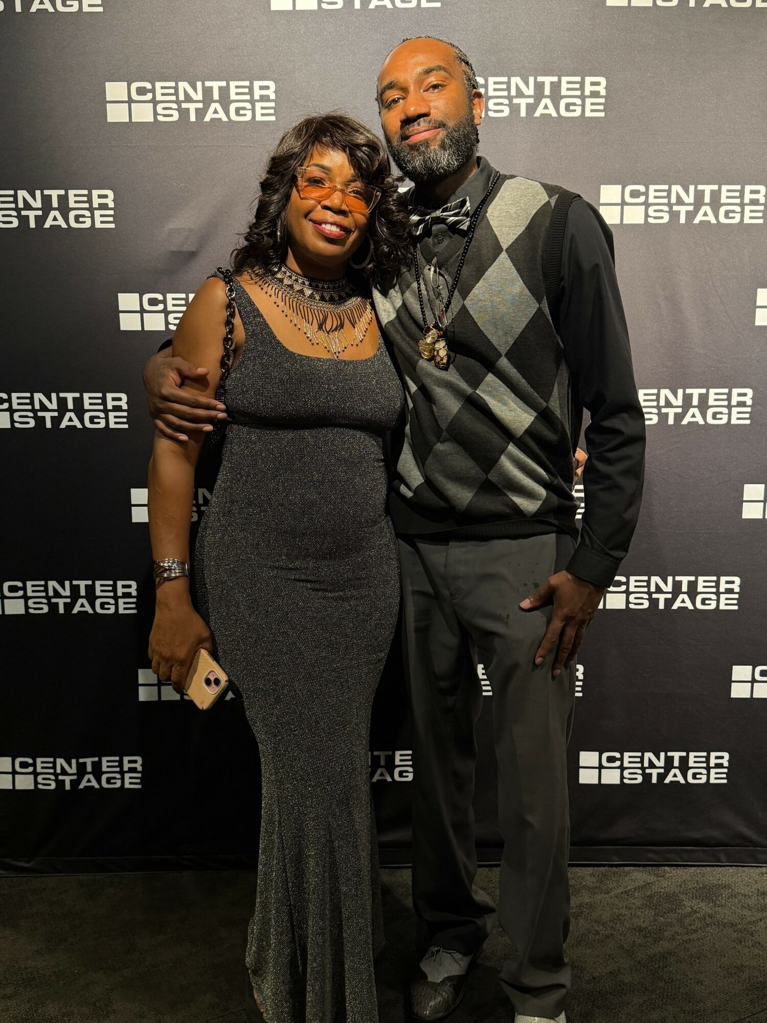 A woman and man pose together in front of a black backdrop with white text, smiling at the camera.