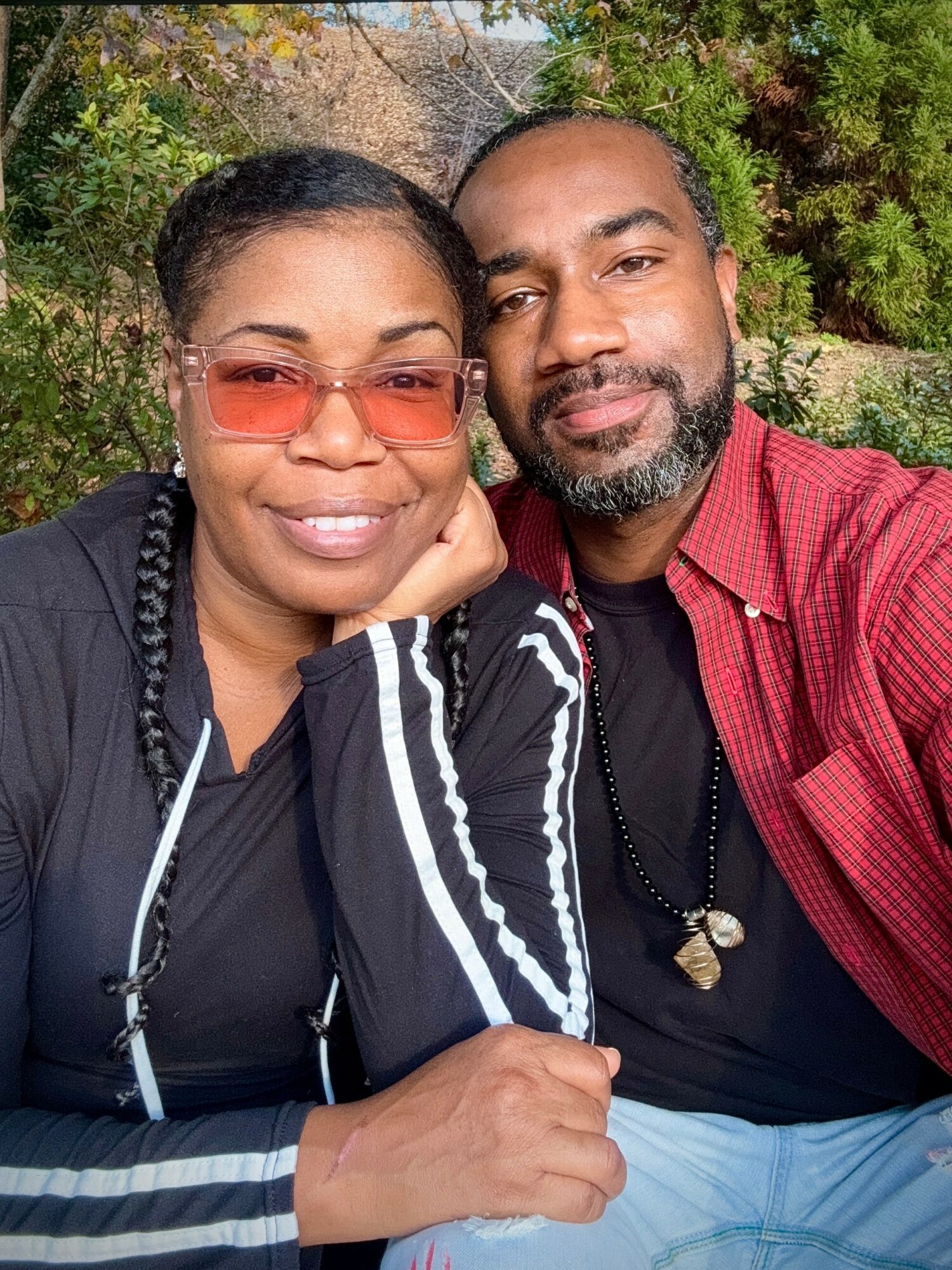 Two people outdoors, smiling, with trees and greenery in background, close-up portrait, wearing casual clothing and glasses.