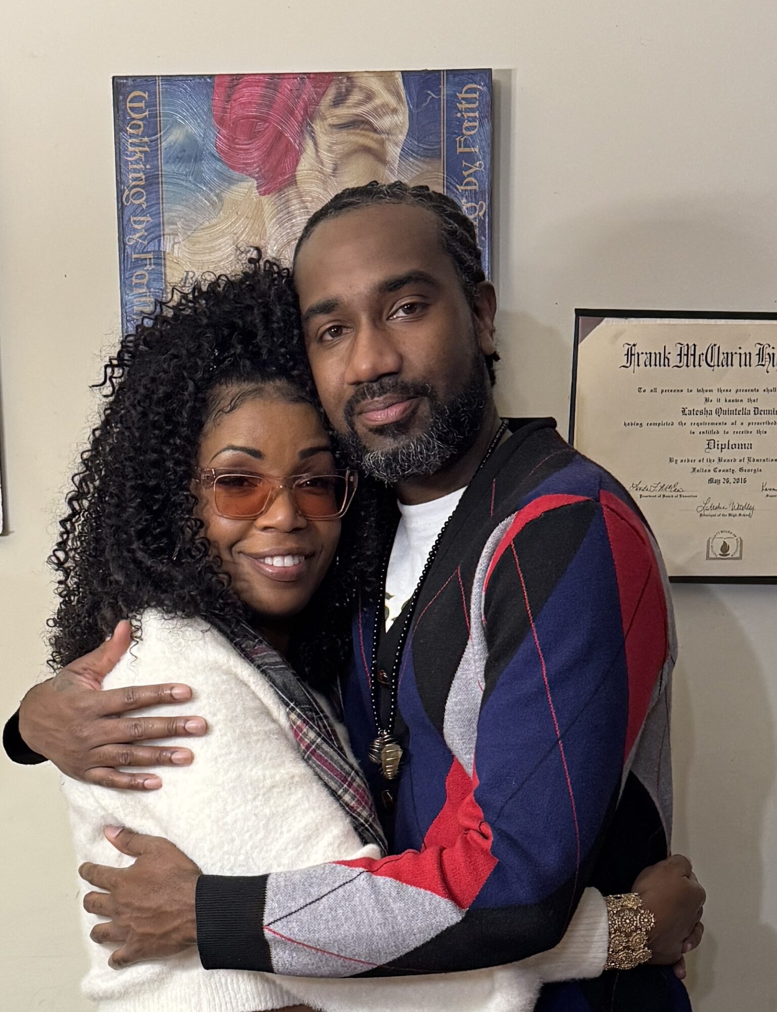 Two people hugging, smiling, in front of framed certificates on a wall, one wearing glasses and a white sweater, the other in a colorful jacket.