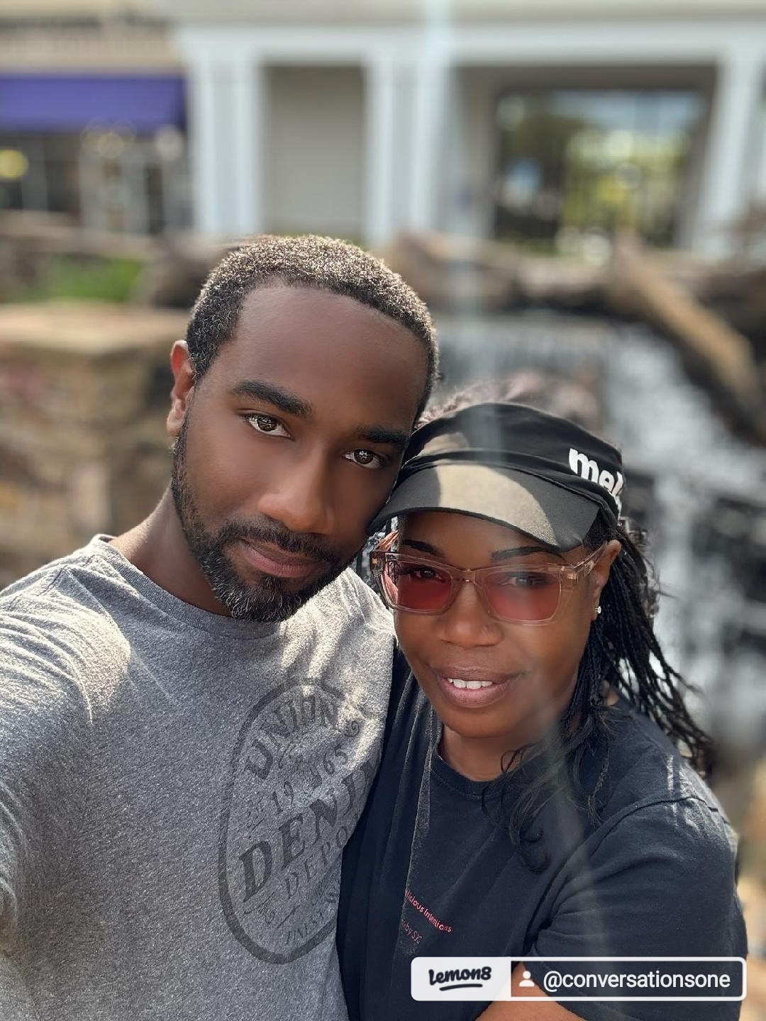 Two people outdoors, one wearing sunglasses and a cap, the other with short hair, smiling at the camera.