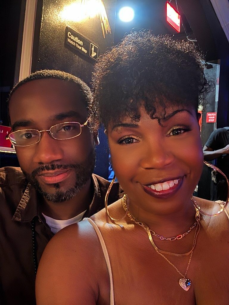 A woman with short curly hair and a man with glasses pose together indoors at night.