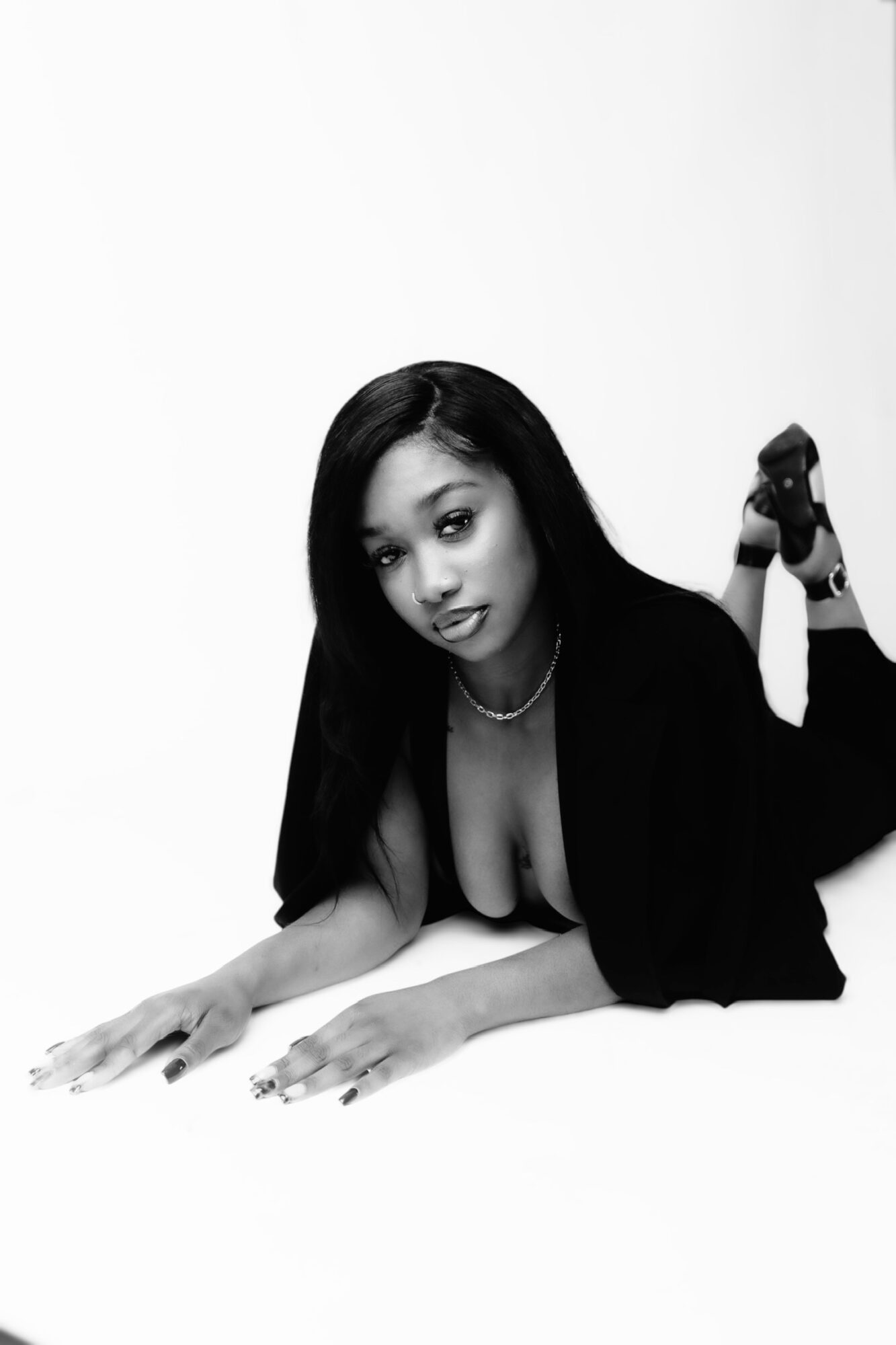 Woman lying on her stomach with legs raised, wearing a blazer and high heels, against a plain background.