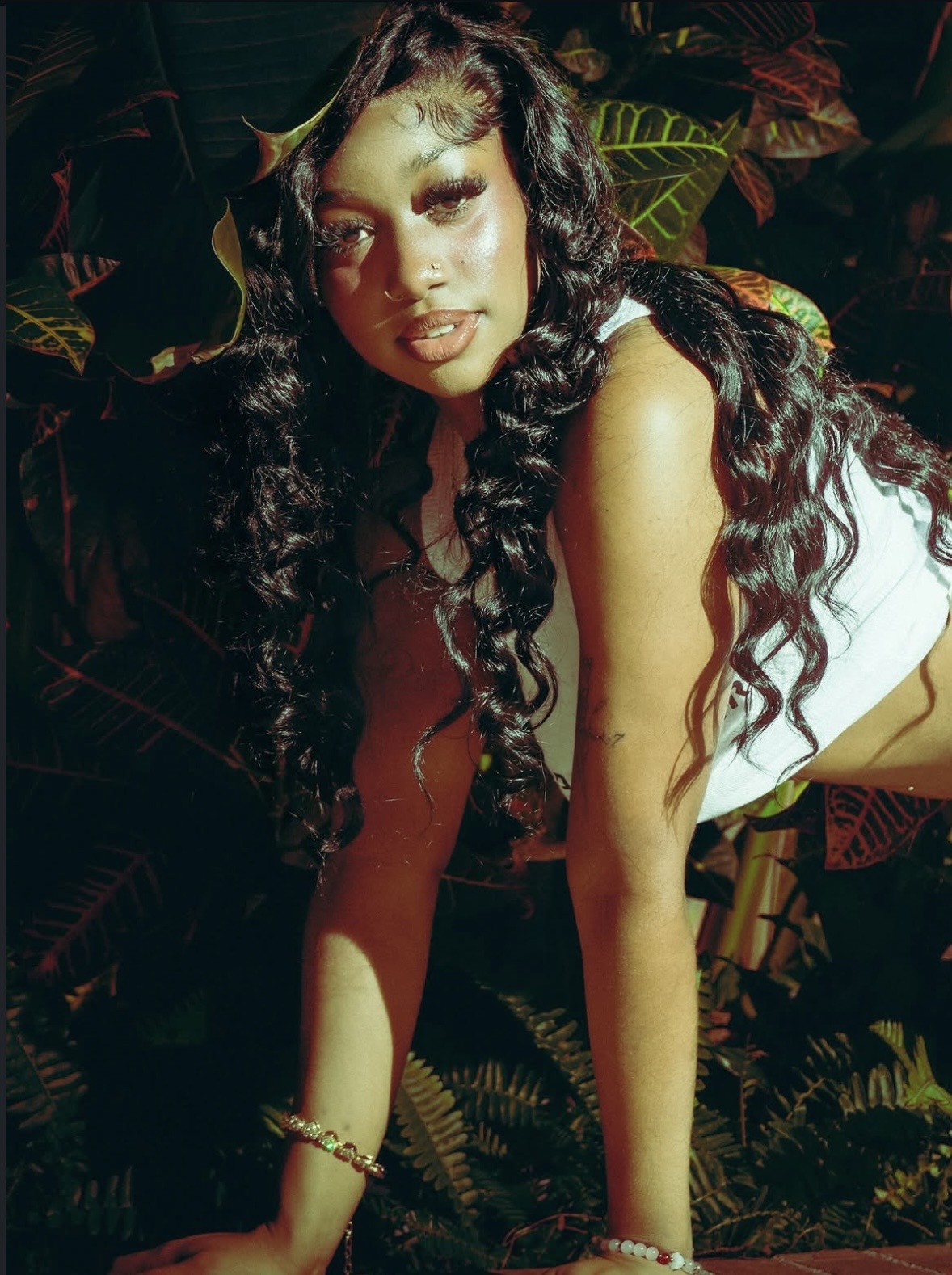 Young woman with long curly hair poses outdoors among tropical plants, wearing a white top and jewelry.