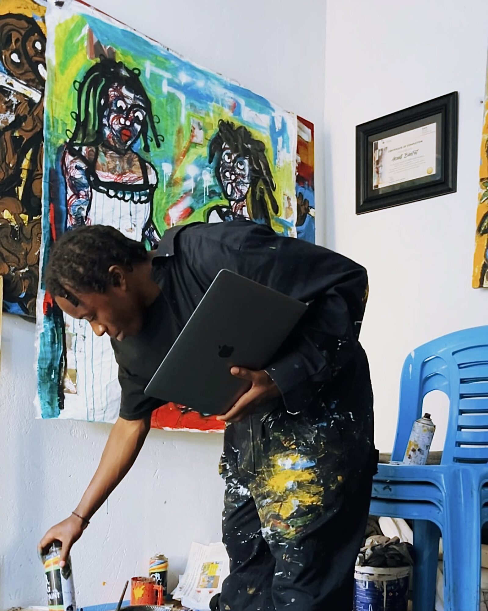 Person with black clothing and a laptop, painting on a wall with colorful artwork, in a room with blue chairs.