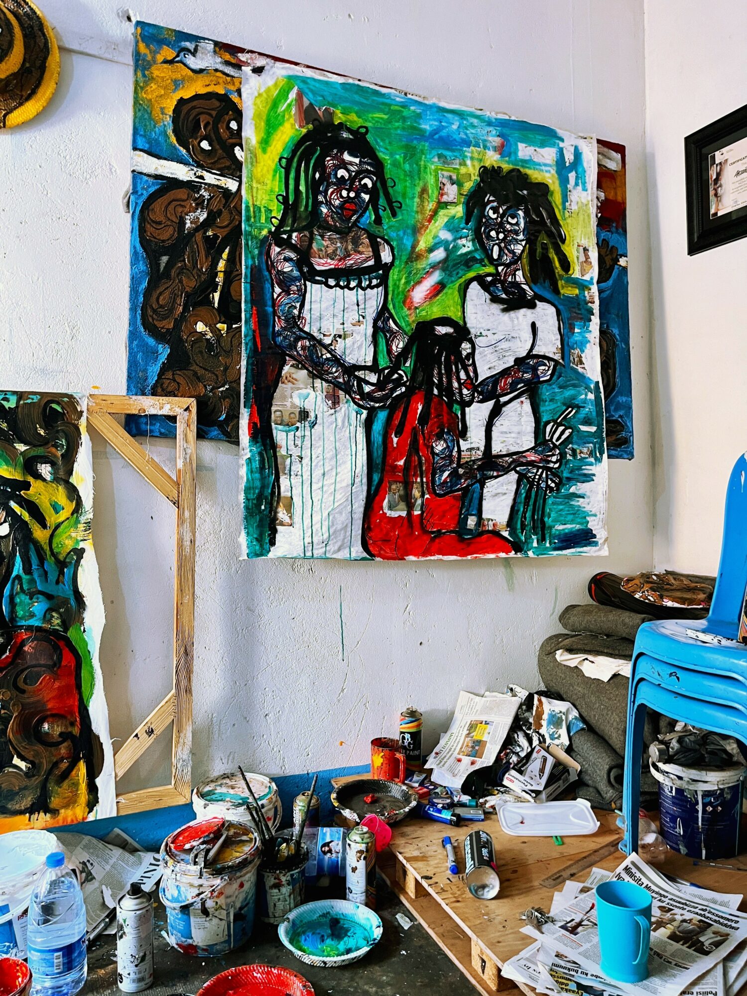 Colorful abstract painting of three figures on wall, with art supplies and furniture in room.