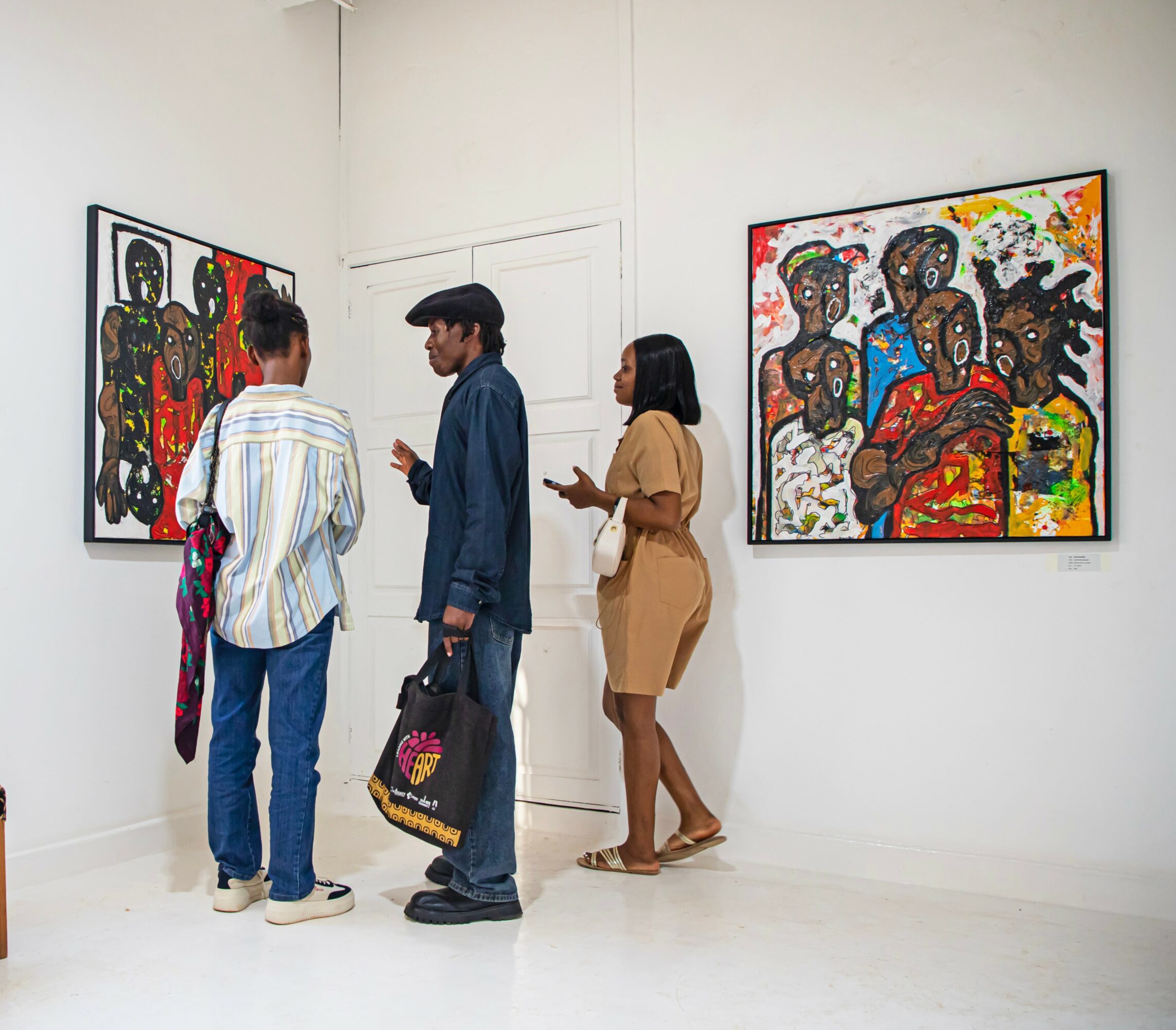 Three people stand in an art gallery, looking at colorful paintings on white walls.