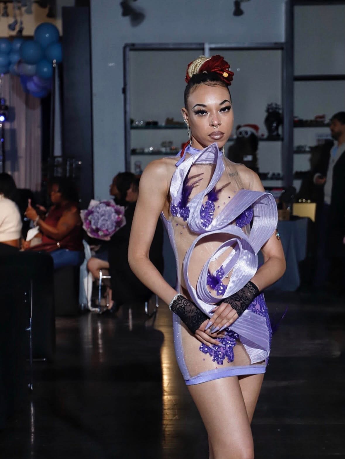 Woman with styled hair and makeup, wearing a purple and silver costume, standing indoors with people and balloons in background.