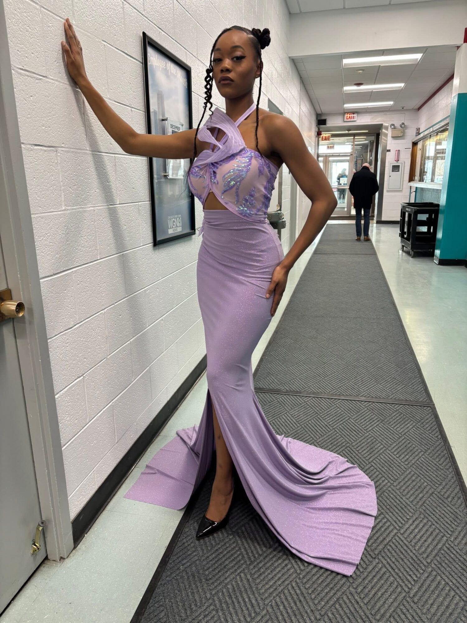 Woman in a lavender gown standing in a hallway, touching the wall with one hand, facing camera.