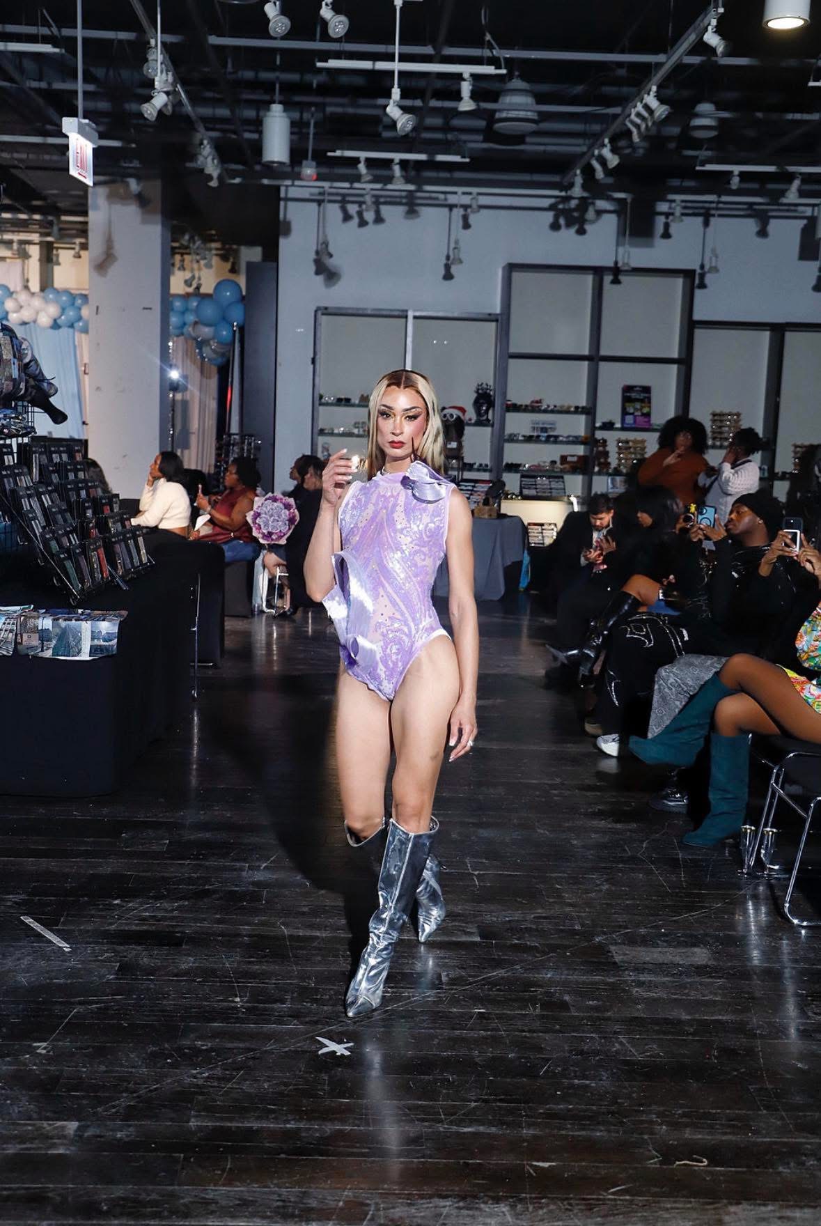 Young woman walking on runway in purple outfit and silver boots, audience seated on both sides, indoor event space.