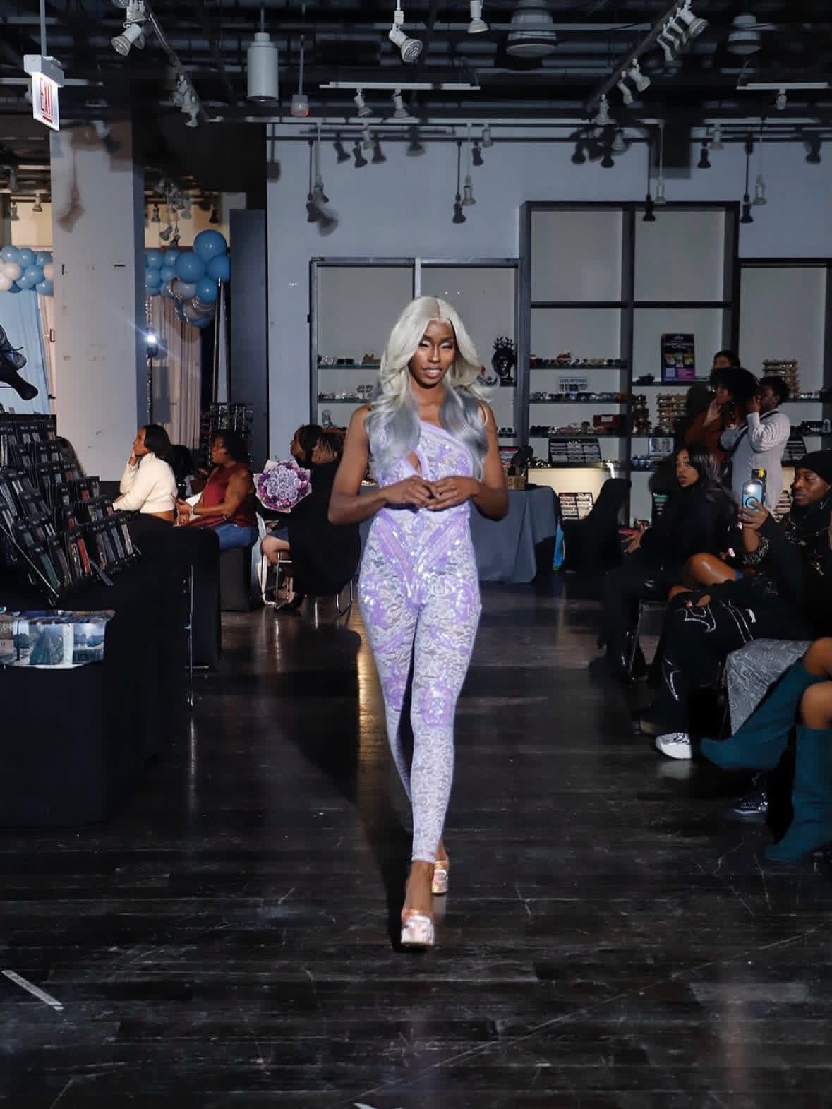 Woman with long silver hair in patterned outfit walking in a modern event space with seated guests and blue balloons.