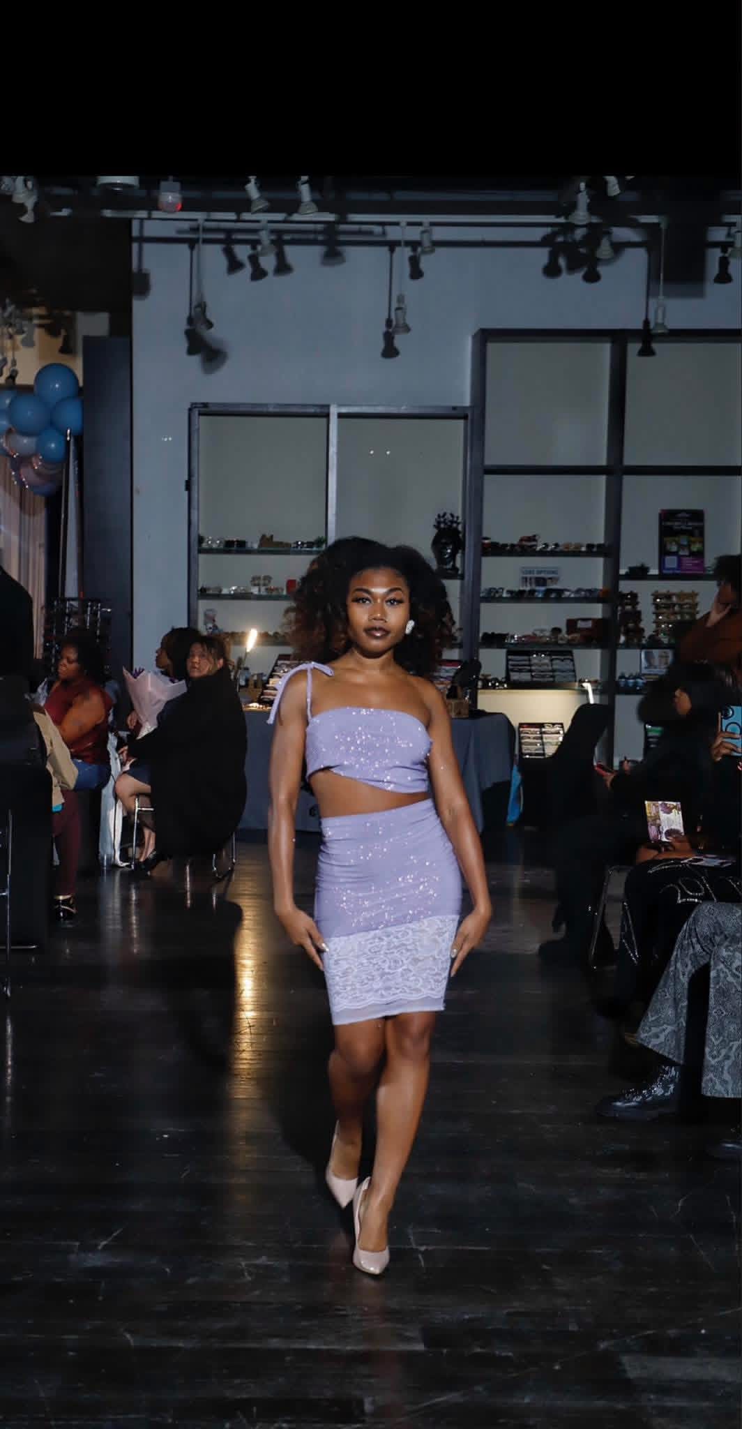 Woman walking on runway in a two-piece outfit, audience seated on sides, industrial-style interior with shelves and lighting.