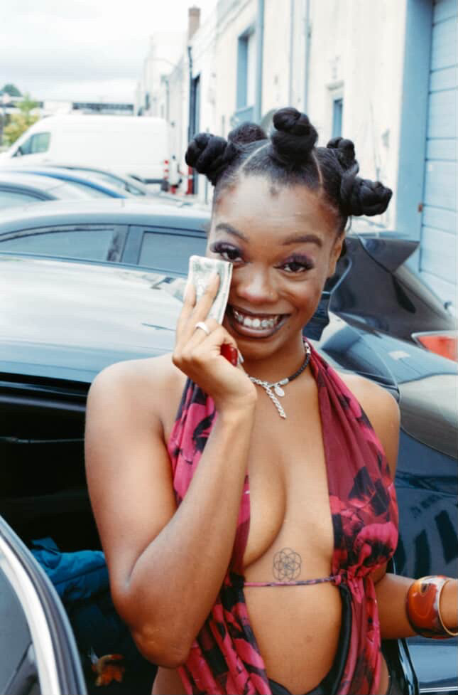 Young woman with styled hair, smiling, holding a dollar bill to her face, wearing a patterned top, outdoors near cars.