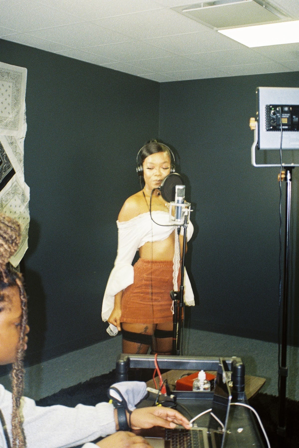 Young woman in a recording studio singing into a microphone with headphones, dark walls, and recording equipment nearby.