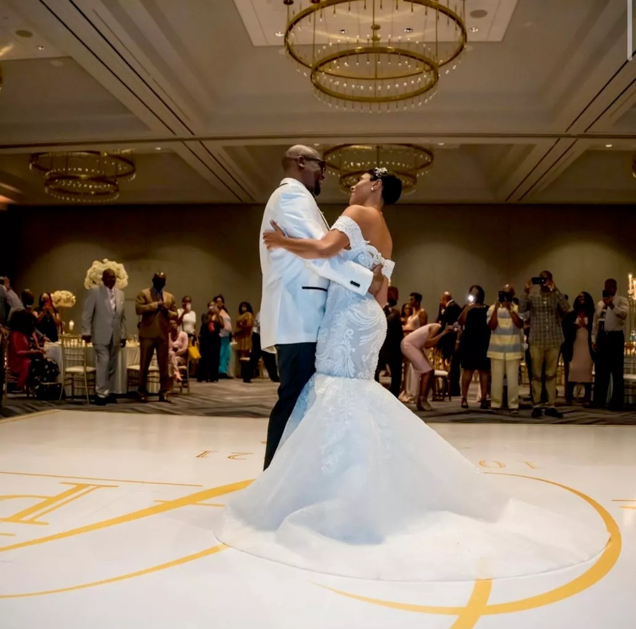 Couple dancing in a ballroom with chandeliers, surrounded by guests, in formal attire.