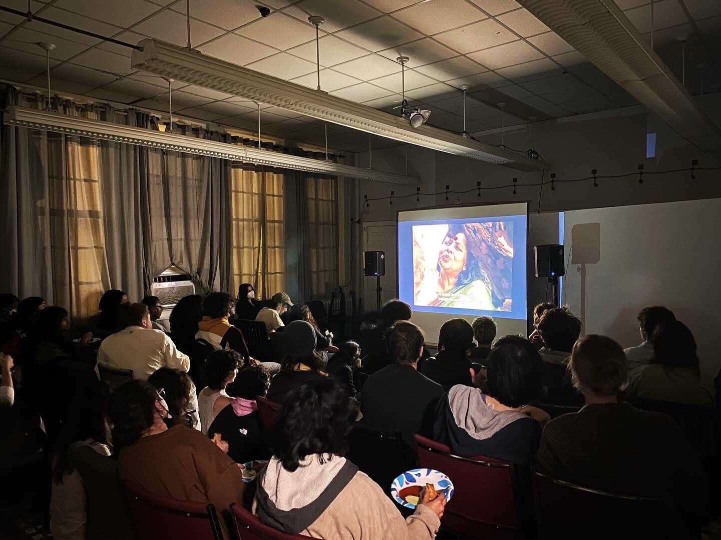 Audience watching a presentation with a large screen showing a colorful image of a person with glasses and curly hair.