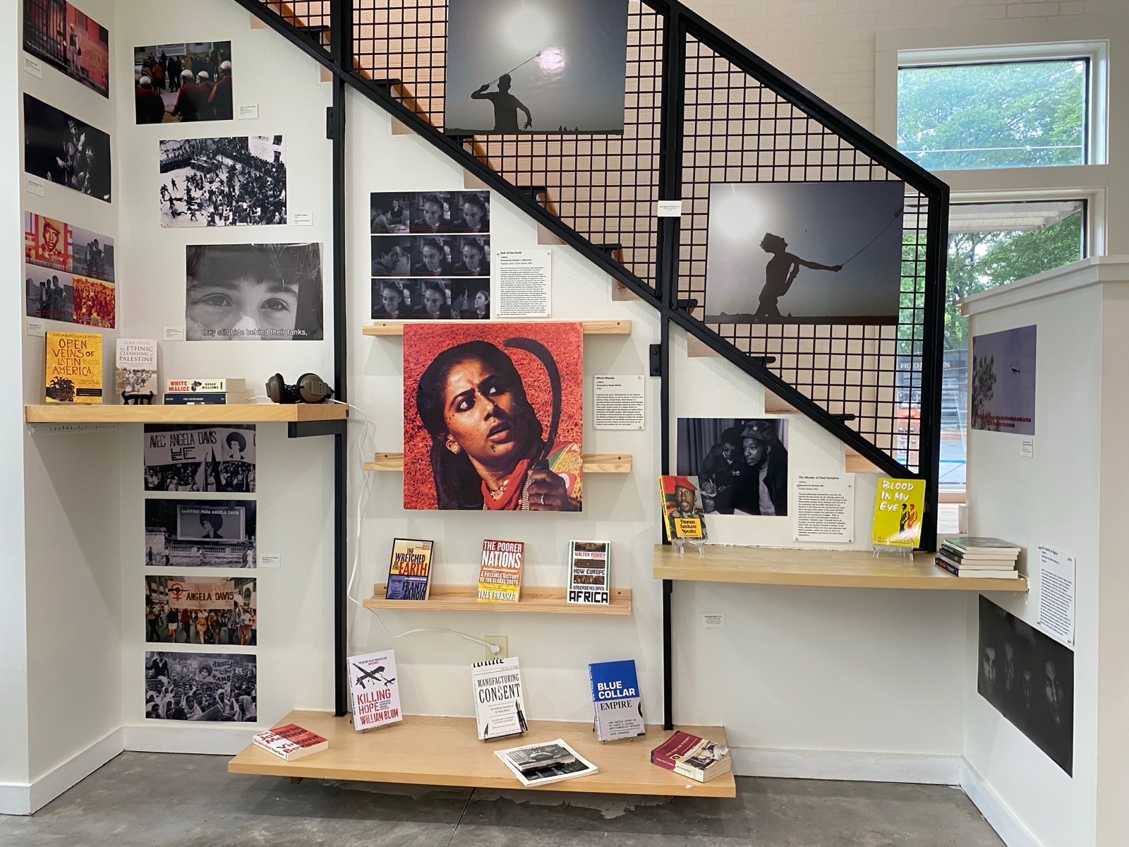 Gallery wall with photographs, artwork, and books, under staircase with a window in background.