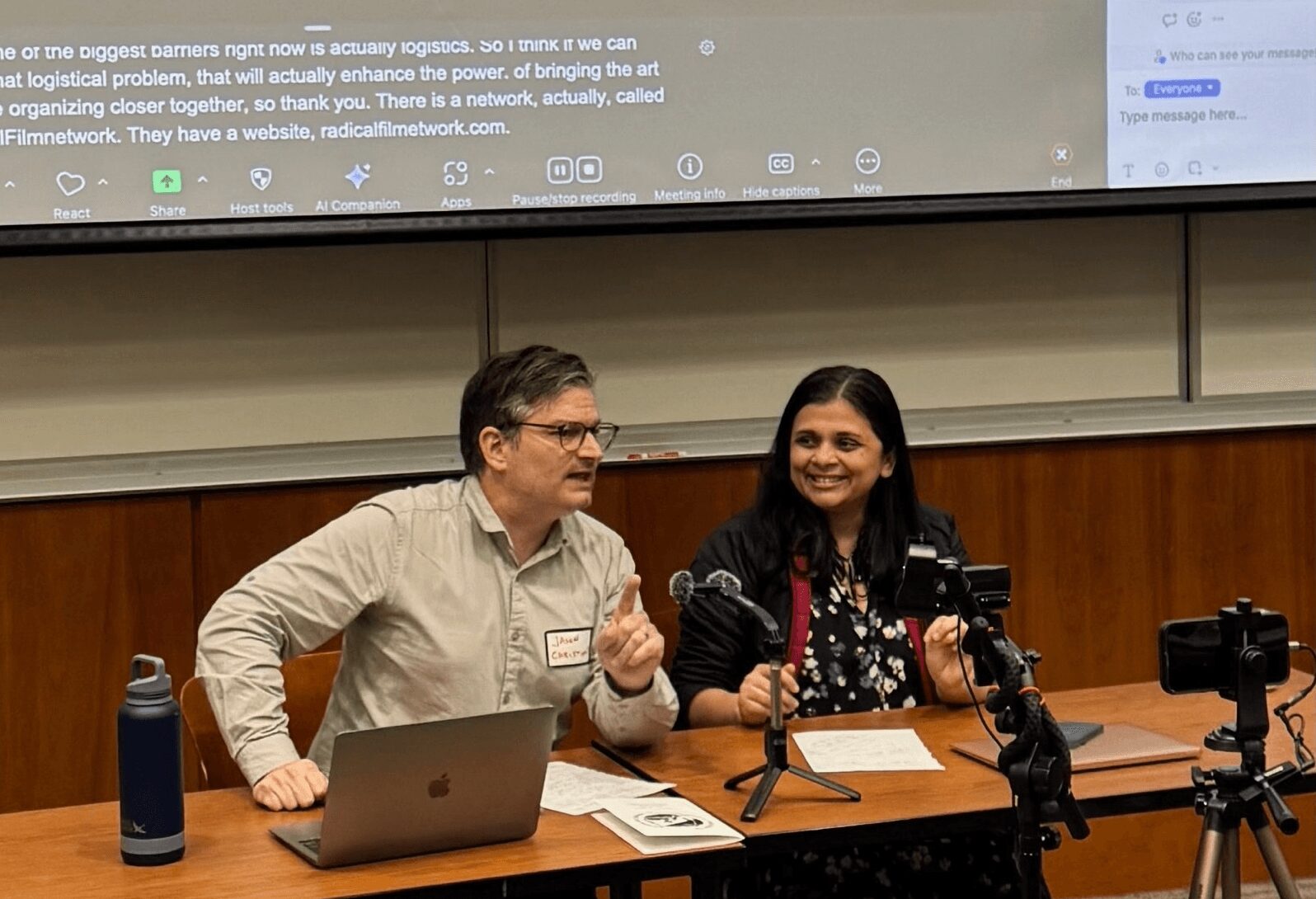 Two people sit at a table with microphones, laptops, and cameras, smiling during a presentation or discussion.