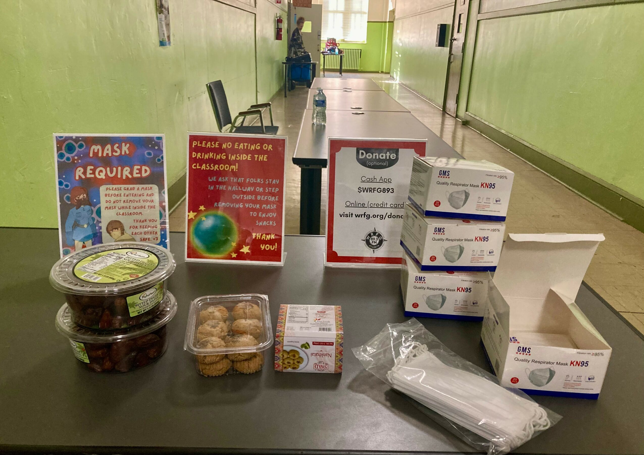 Table with boxes of masks, hand sanitizer, and snacks in a hallway with green walls.