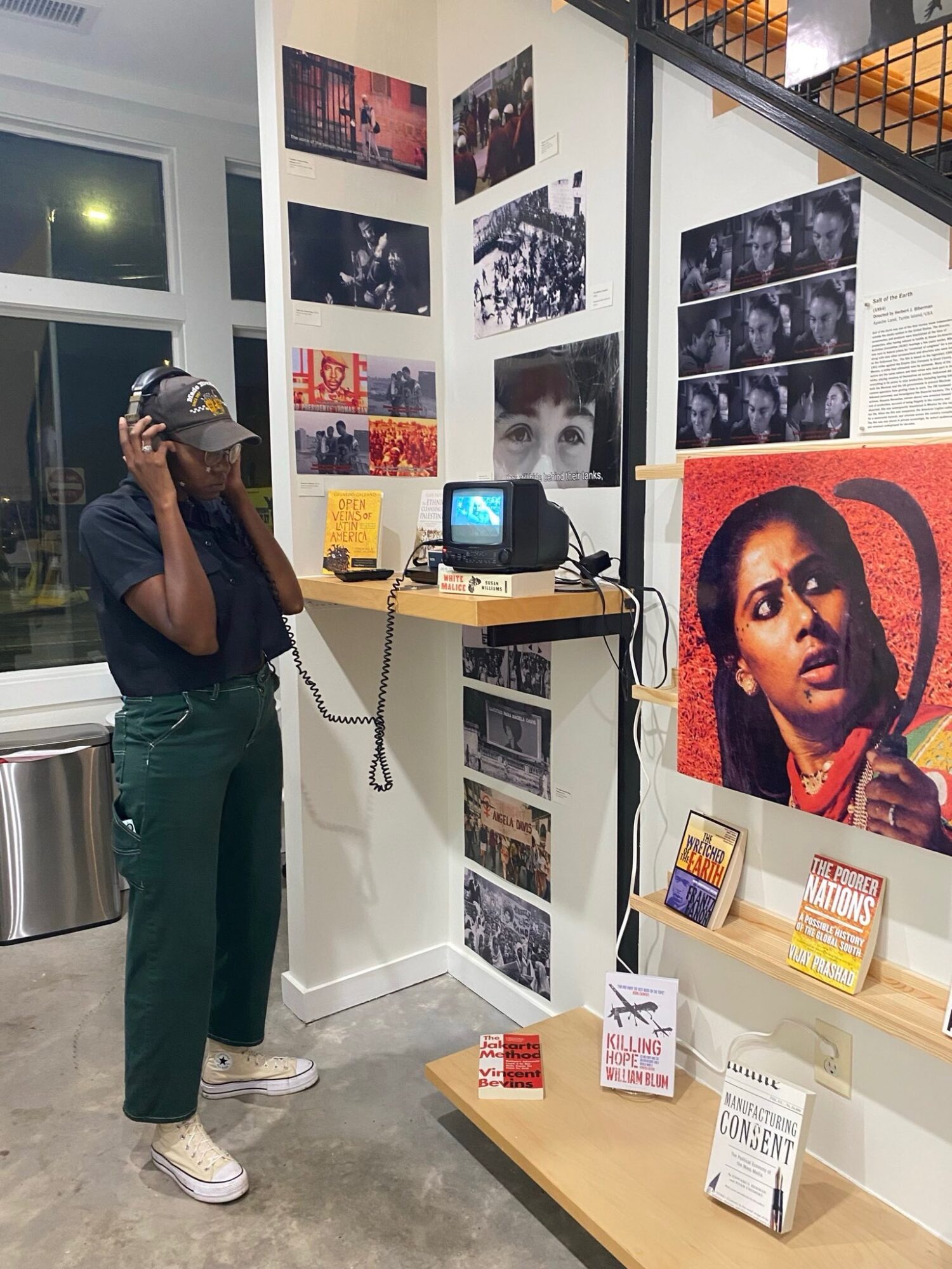 Woman in dark shirt, green pants, and cap using a vintage telephone in a gallery with photographs and artwork displayed on white walls.