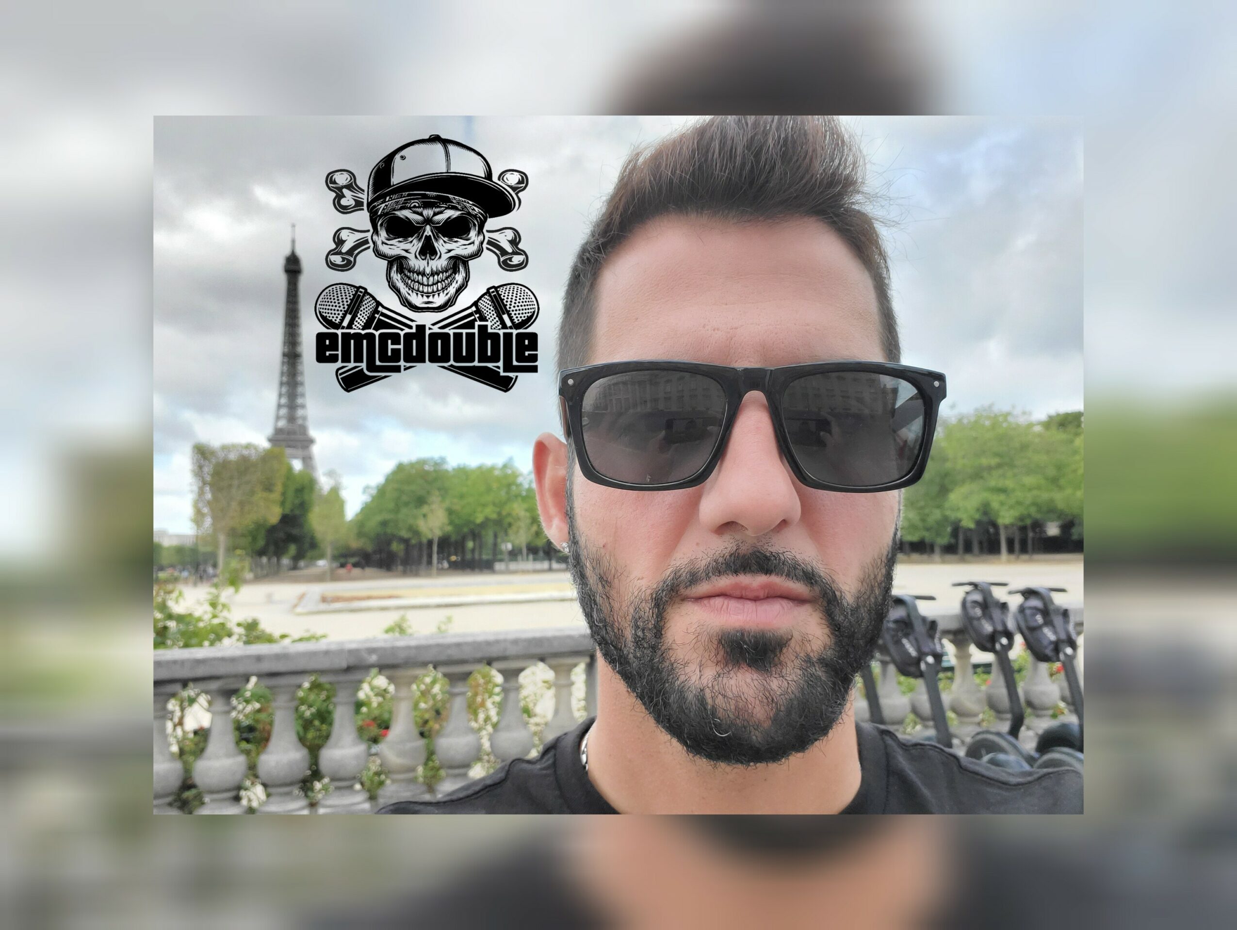 Man with sunglasses and beard outdoors with trees and the Eiffel Tower in the background.