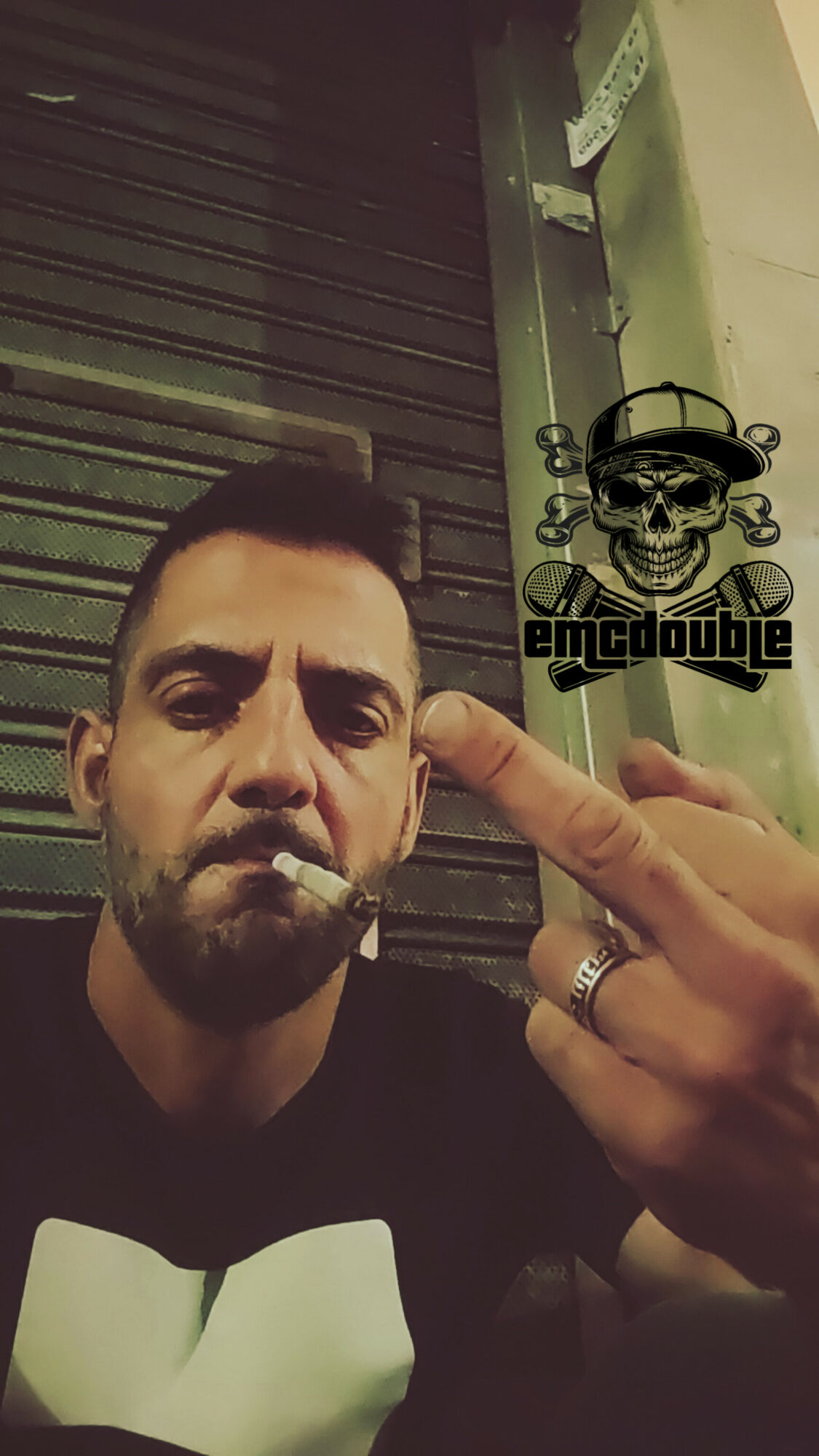 Man with a cigarette making a rude gesture, holding up his middle finger, in front of a dark background.