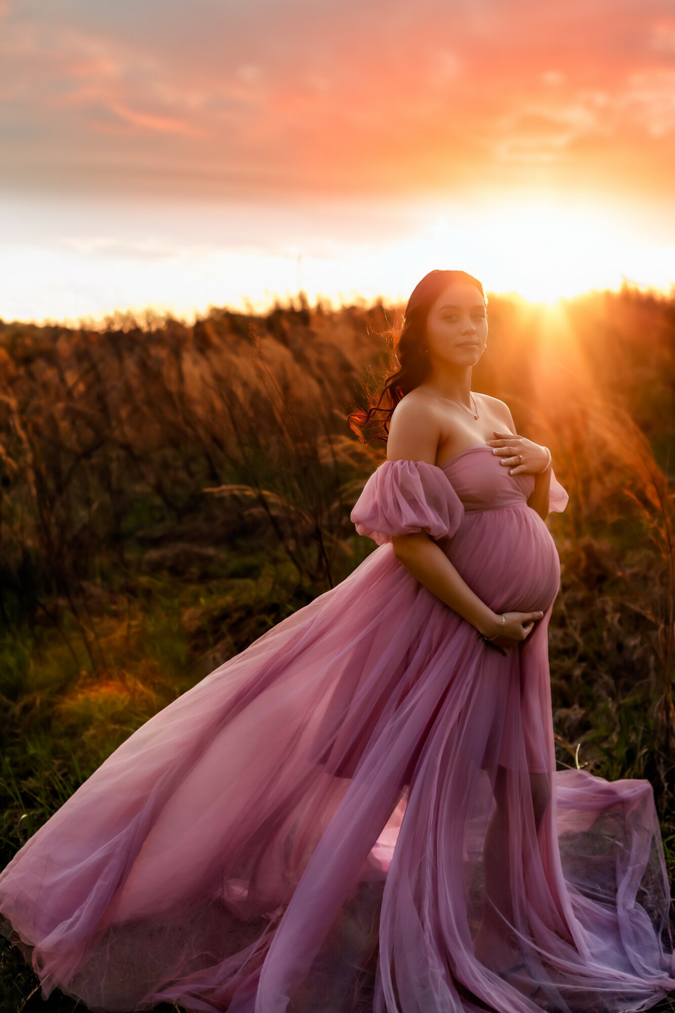 Pregnant woman in a flowing pink dress stands outdoors during sunset, holding her belly with both hands.