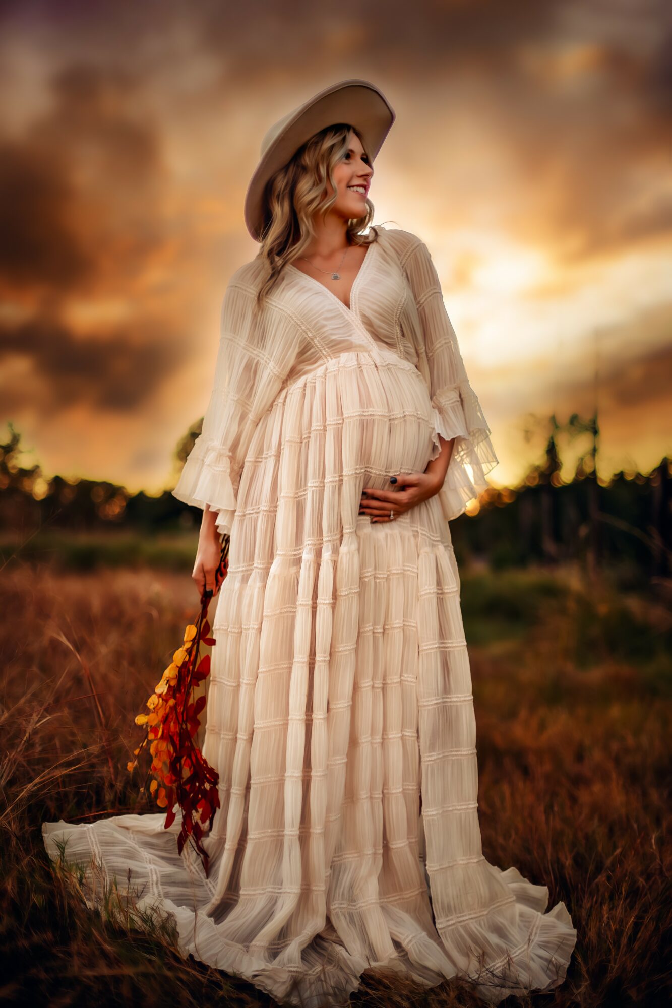 Woman in a flowing dress and wide-brimmed hat standing outdoors at sunset, holding a bouquet of flowers.