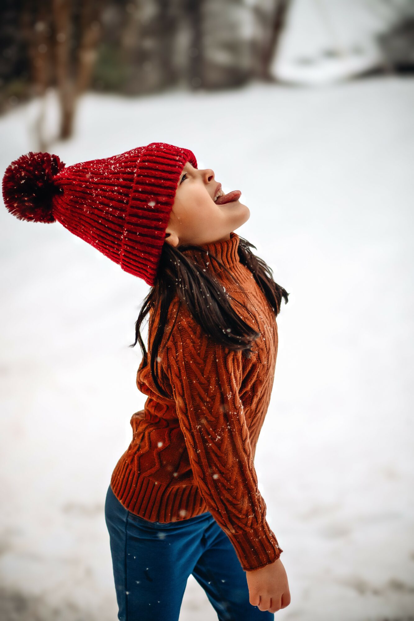 Girl in winter clothing with red knit hat and orange sweater standing outdoors in snow.