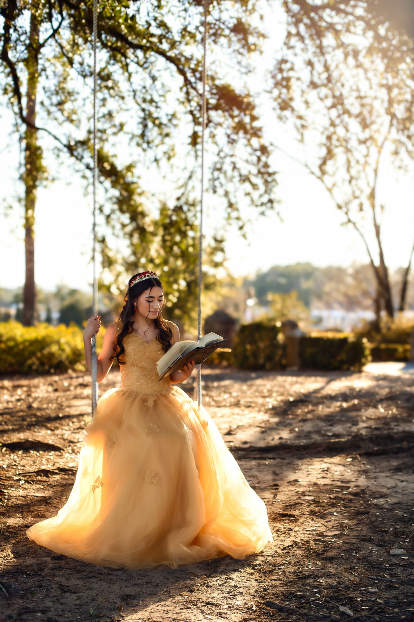 Young woman in a yellow dress sitting on the ground reading a book outdoors with trees and sunlight.