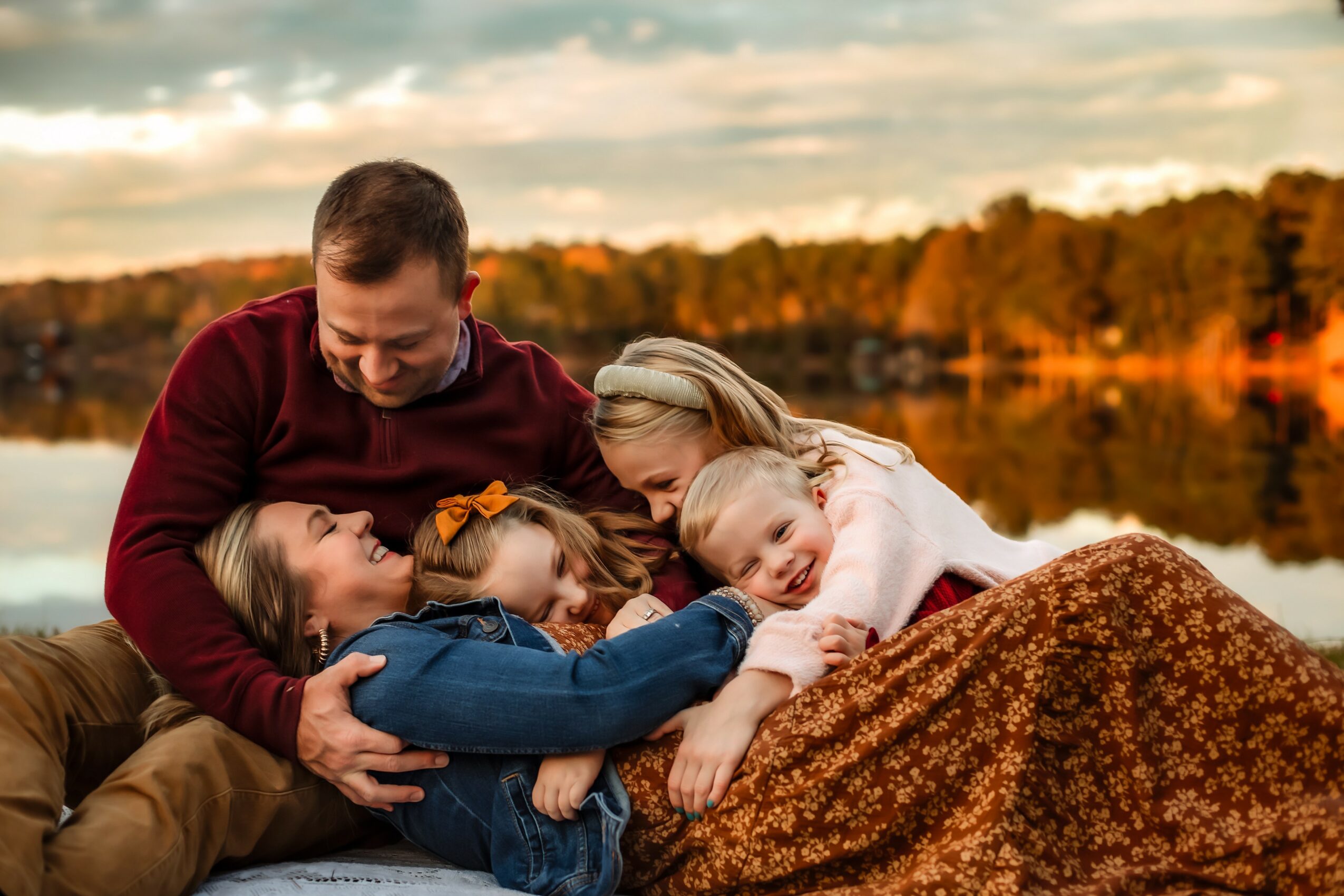 Family of four lying outdoors near a lake, smiling and cuddling together during sunset.