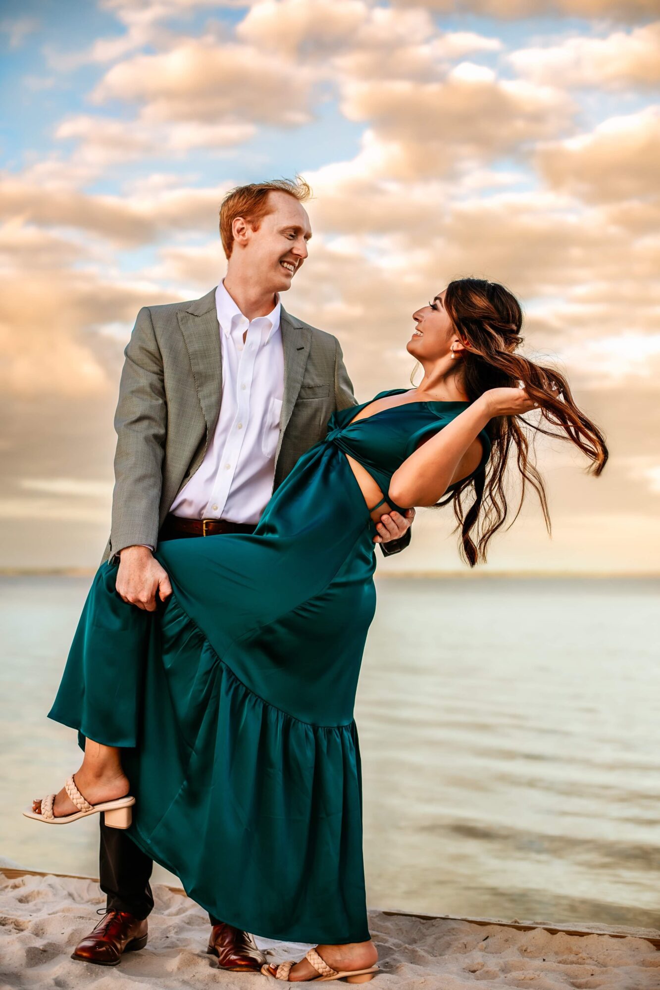 Man in gray suit holding woman in teal dress by the water, sky with clouds in background.
