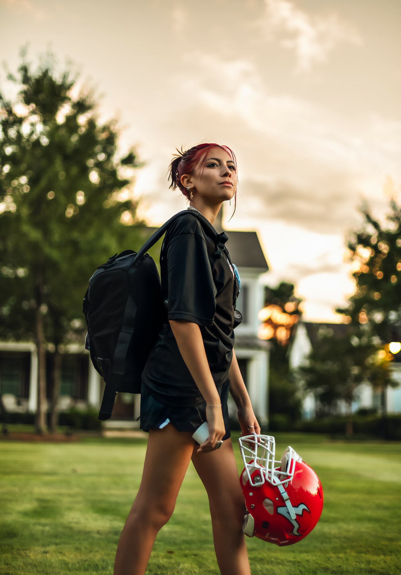 Young woman with a backpack holding a helmet, standing outdoors on grass with trees and buildings in background.