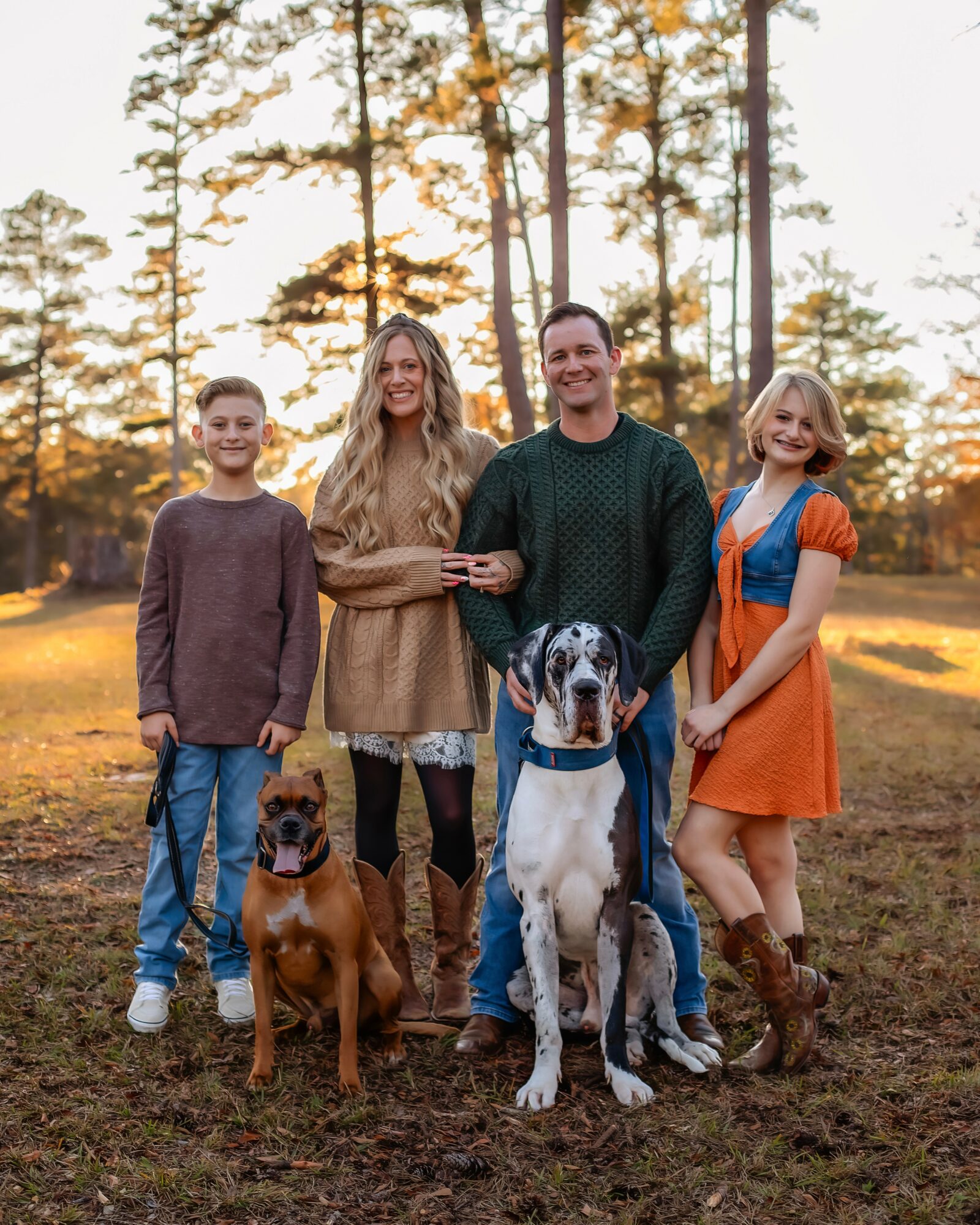 Family of four with two dogs standing outdoors in a park during sunset, trees in background, smiling faces.