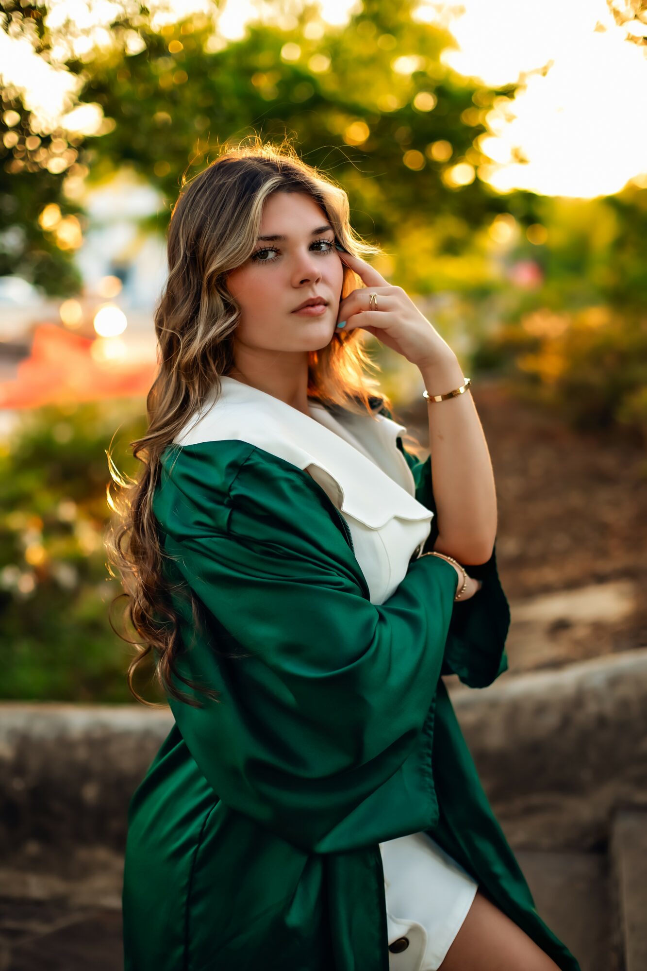 Young woman with long wavy hair in a green graduation gown outdoors, touching her face, with trees and sunlight behind her.