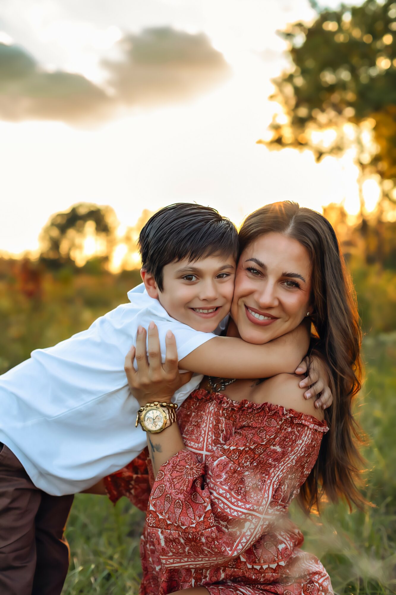 A woman and a young boy outdoors at sunset, smiling, with trees and sky in background.