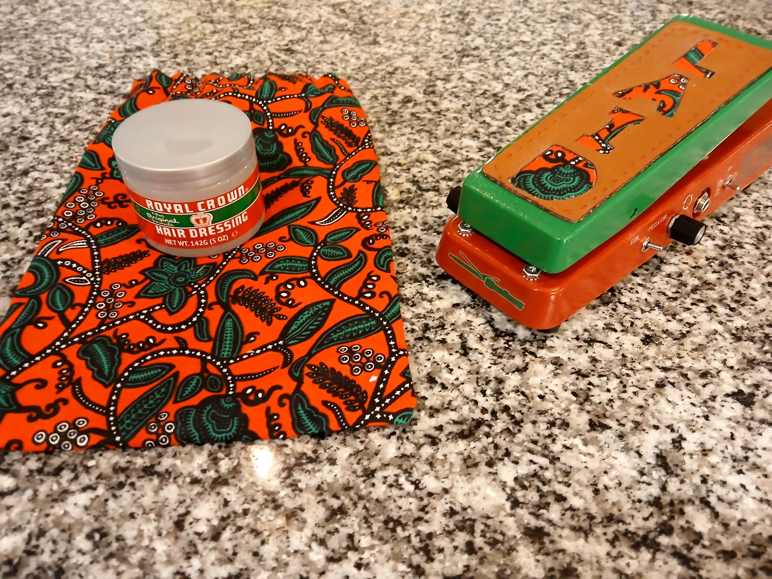 A small container with a white lid, a colorful fabric pouch, and a decorated box on a speckled countertop.
