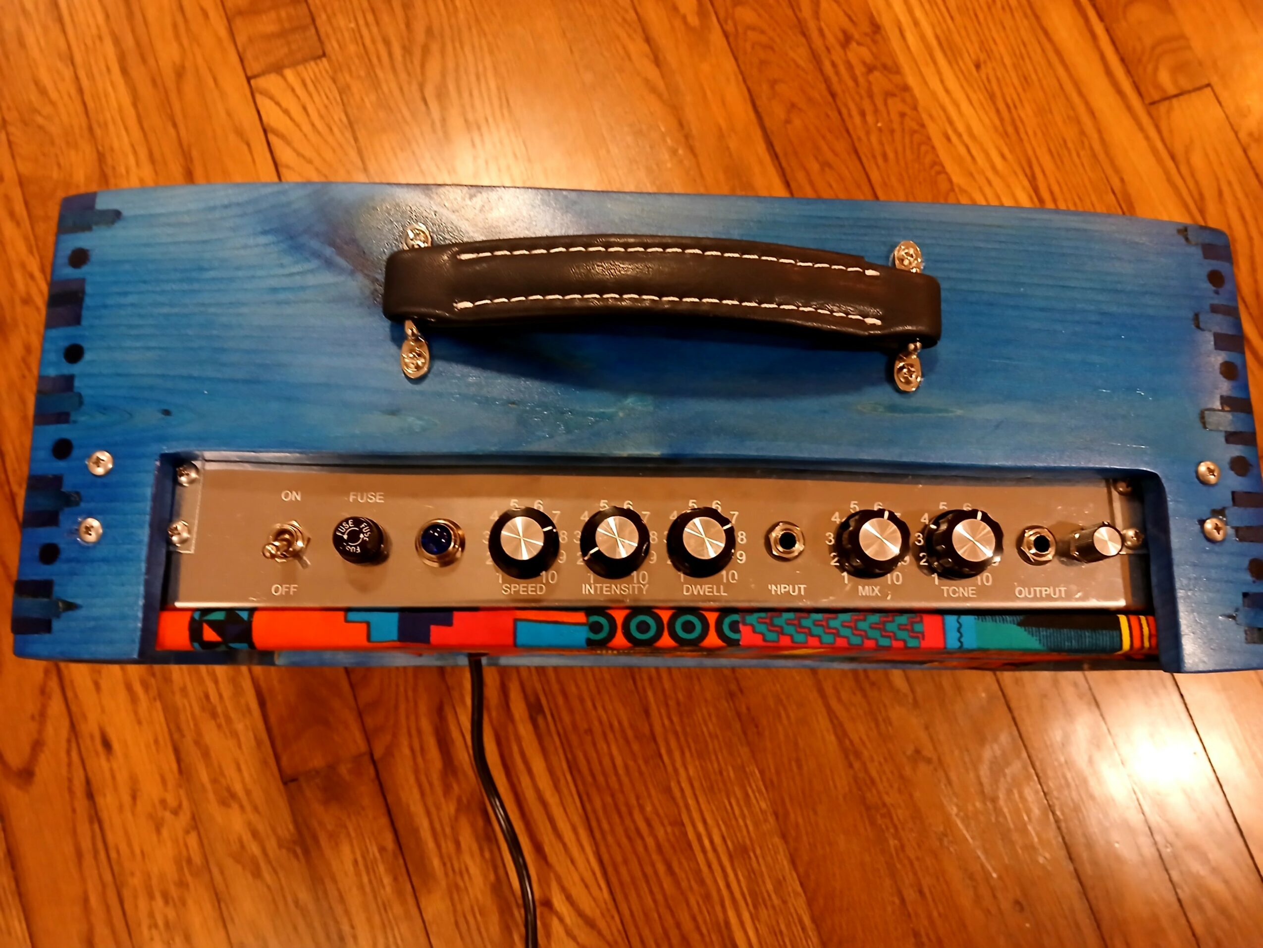 Blue wooden object with a black handle on top, metal control panel with knobs, and colorful markings, on a wooden floor.