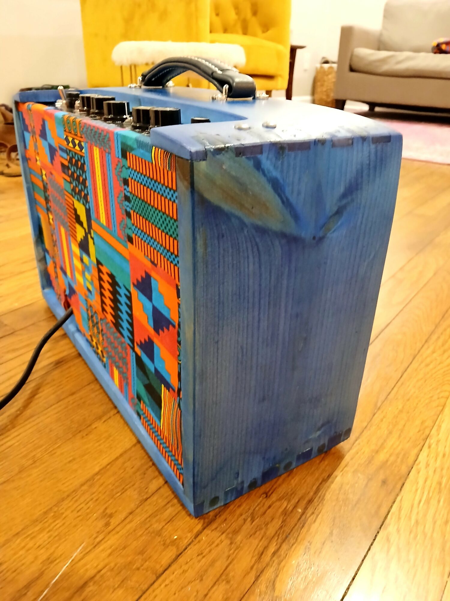 Box with colorful geometric patterns on side, blue painted top, on wooden floor, in a living room with yellow and beige furniture.