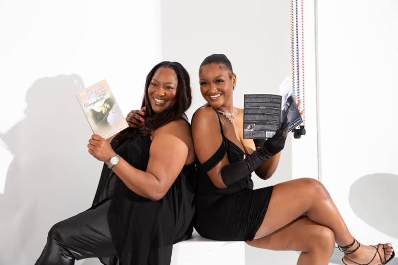 Two women in black dresses and gloves sit back-to-back, smiling and holding books in a bright room.