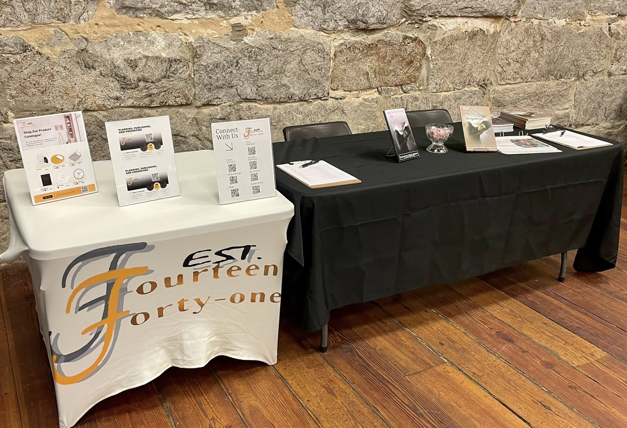 Two tables with promotional materials and displays, one covered with a white cloth and the other with a black cloth, against a brick wall.