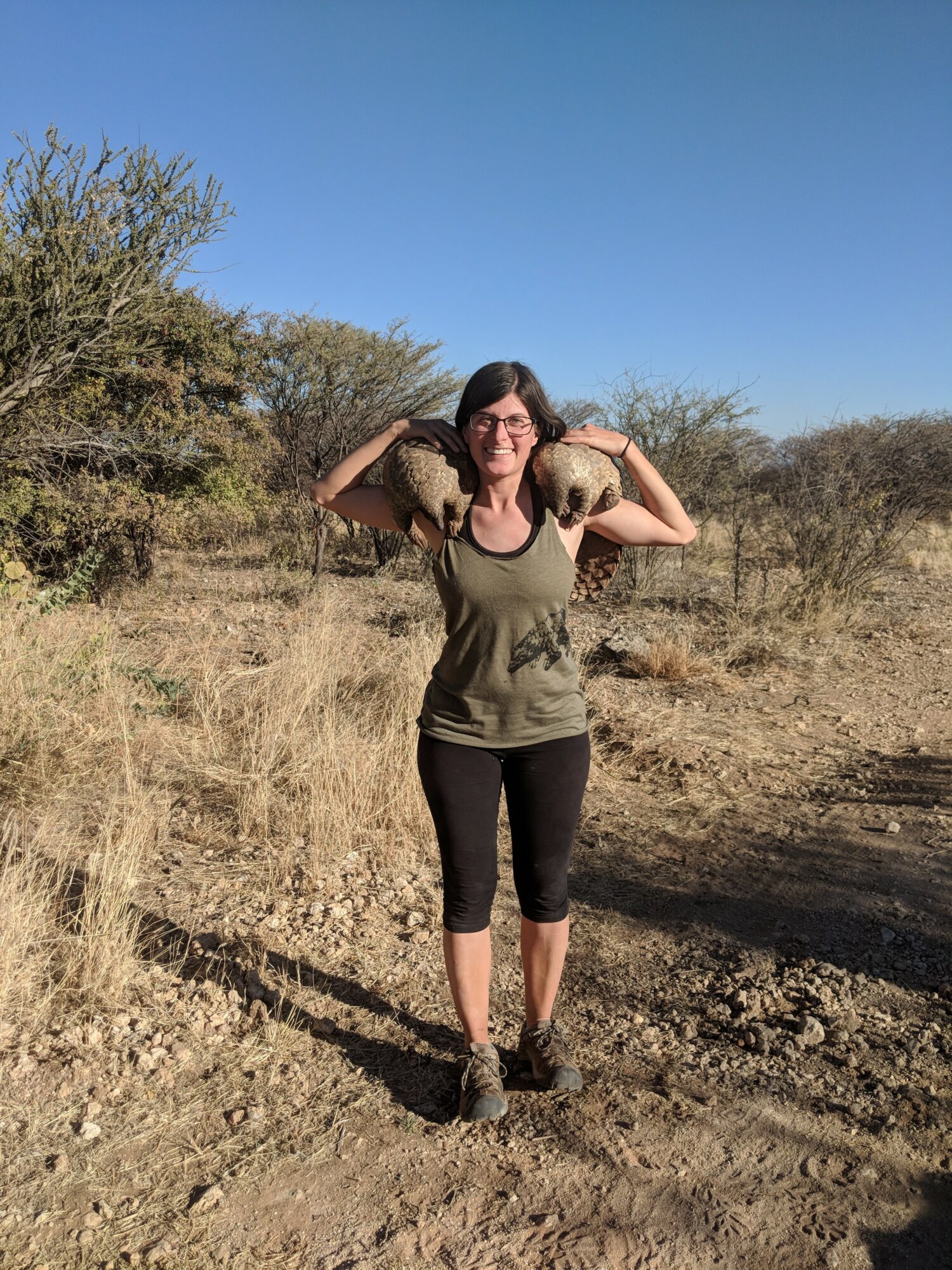 Woman standing outdoors on dirt path, holding two large rocks on her shoulders, with bushes and clear blue sky behind her.