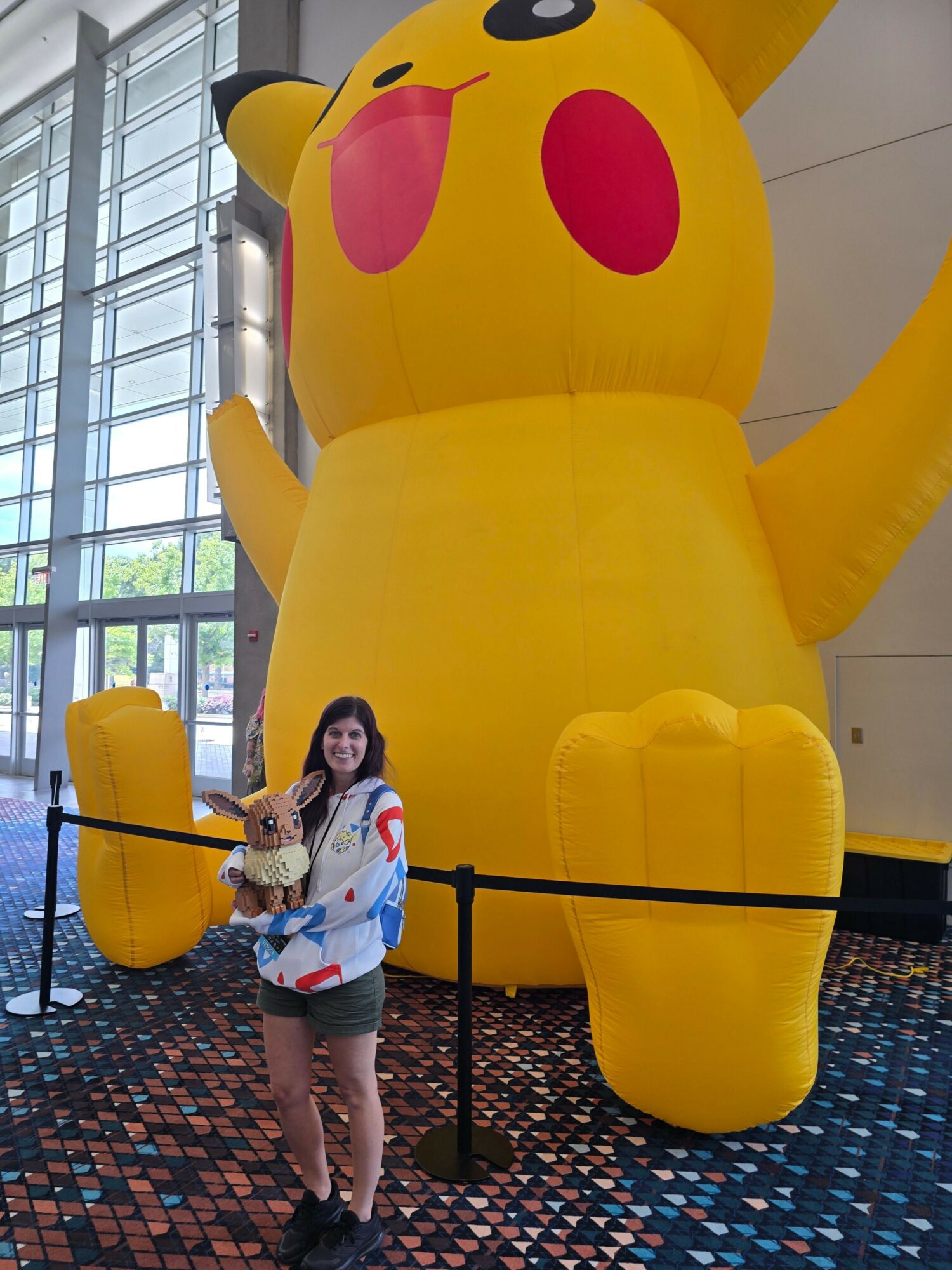 Person standing in front of a large Pikachu inflatable with a plush toy, inside a building with glass windows.