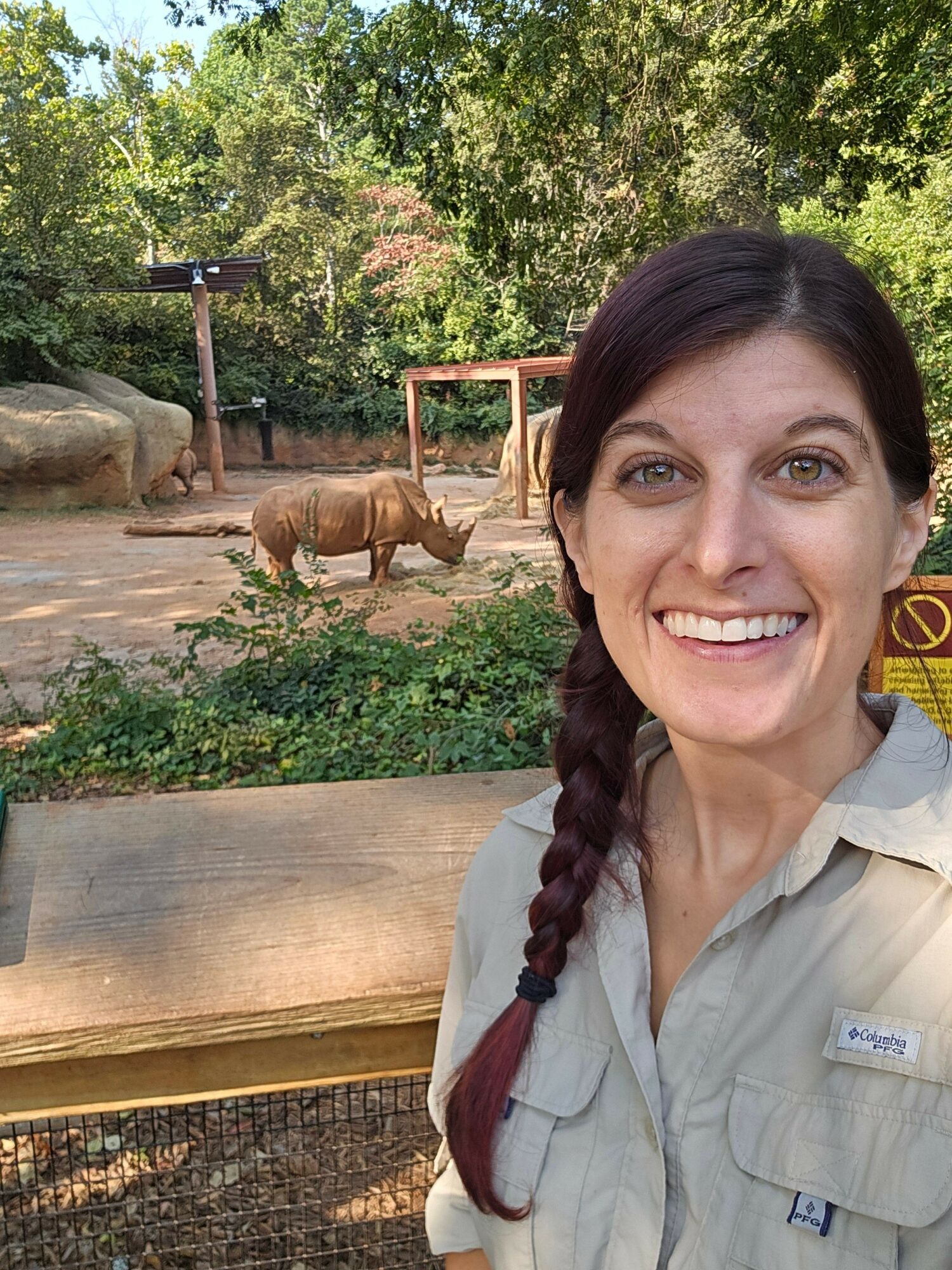 Woman with dark hair in braid smiling outdoors, with a rhinoceros in the background among trees and rocks.