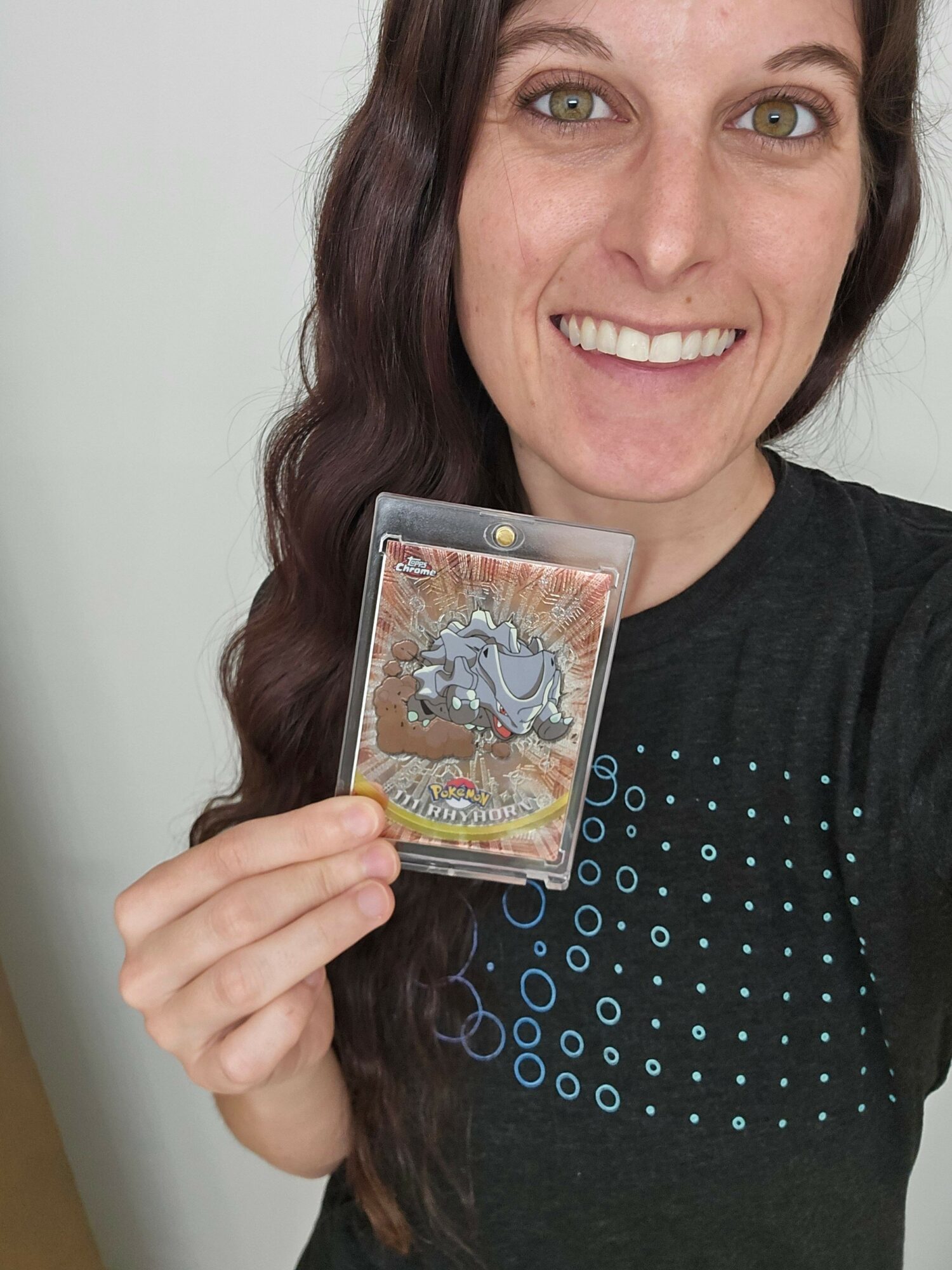 Woman with long dark hair smiling, holding a card with a dinosaur illustration, wearing a black shirt with blue dots.