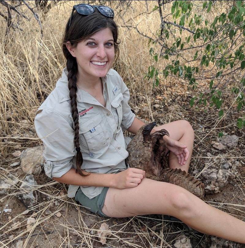 Young woman with long braided hair sitting outdoors on dry grass, smiling, holding a small animal.